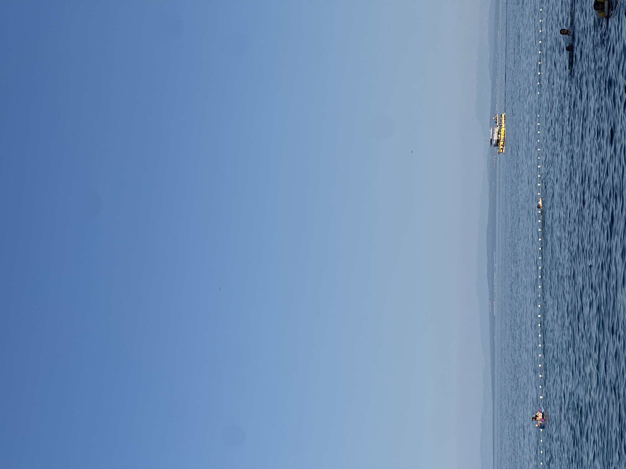 The image depicts a serene view of the Adriatic Sea near Lovran, Croatia. The sky is a clear, bright blue with a few distant, faint smudges. The sea is a deep blue with gentle ripples. A white buoy line stretches across the water, marking a swimming area.
In the distance, a yellow and white tourist boat with multiple decks is cruising across the sea. On the left side of the frame, a person is relaxing on an inflatable tube within the marked swimming area. Further into the water, several other individuals are visible swimming, with some partially submerged. The distant horizon shows a faint outline of a coastline with hills or mountains, suggesting the presence of land beyond the sea. The overall atmosphere is calm and recreational, typical of a summer day at the seaside.