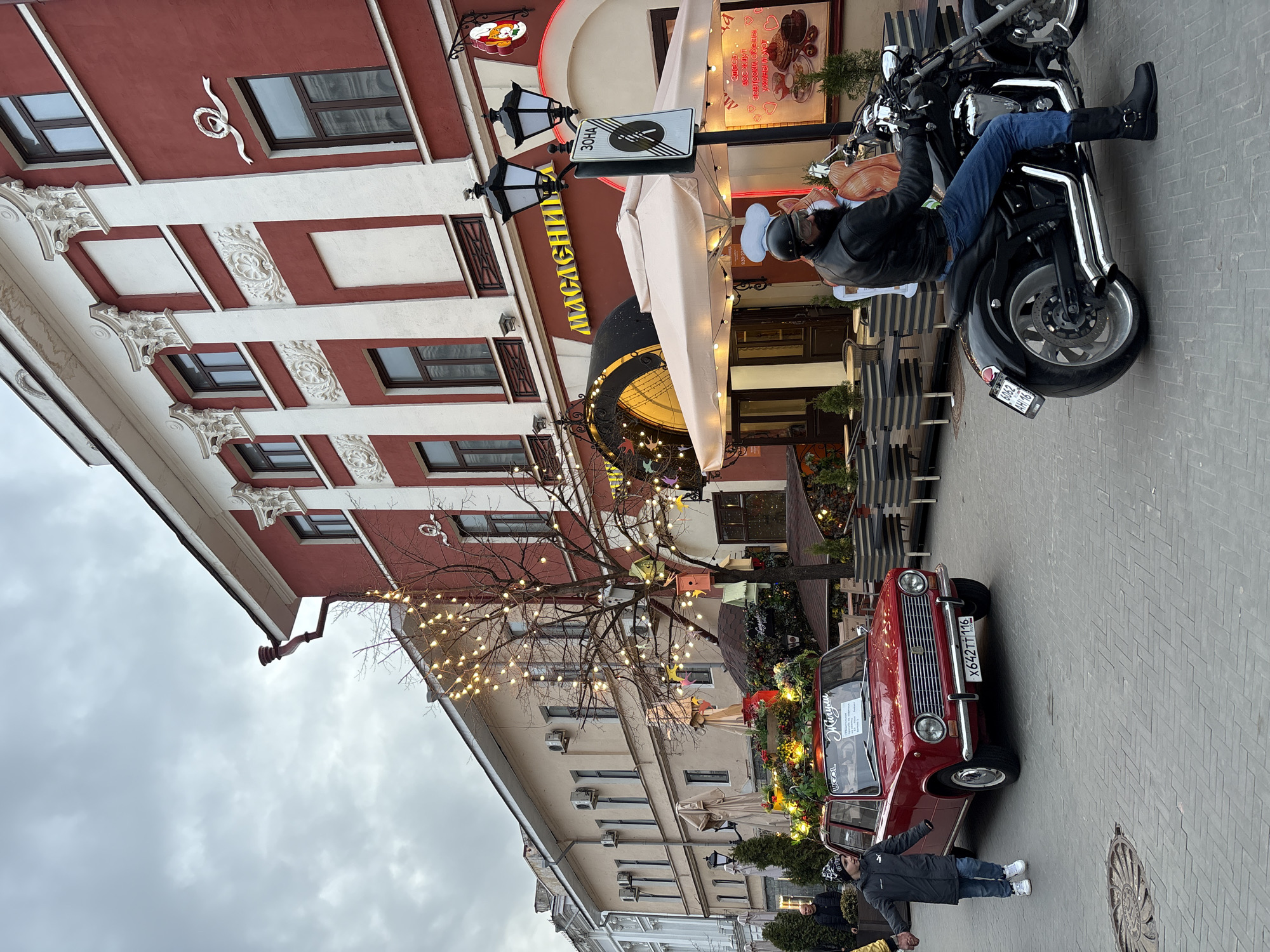 This image captures a lively street scene in Kazan, Russia, under an overcast sky. In the foreground, a man in a black leather jacket, helmet, jeans, and boots poses on a black motorcycle parked on the paved sidewalk. Nearby, another man in a dark jacket, jeans, and white sneakers stands with outstretched arms next to a red vintage-style car, identified as a "Жигули" (Zhiguli) by text on its windshield, also decorated with festive elements and "UGOL". Both vehicles display Russian license plates: "X642 TT 116" on the car and "6062 AH 16" on the motorcycle.
The background is dominated by a reddish-brown building featuring ornate white architectural details, including decorative cornices and panels. This building houses a cafe or restaurant, indicated by a large yellow sign reading "МАСЛЕНИ" (Maslenitsa) and another sign advertising "завтраки, обеды, ужины и уют" (breakfasts, lunches, dinners, and coziness). Outdoor dining tables and chairs are visible under beige awnings, with bare trees strung with decorative lights adding to the ambiance. A street sign nearby marks a "ЗОНА" (Zone) with a "no outside food/drink" symbol. The scene combines urban life with festive decorations and classic vehicles.