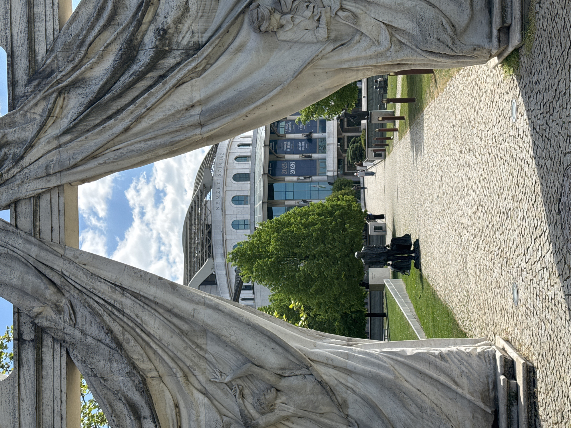 An outdoor scene captured during the daytime shows a view through a large, artistic archway made of draped, weathered stone sculptures, likely part of a monument. The sculptures, which appear to include a human figure integrated into the drapery on the left, frame a paved pathway leading towards a prominent modern building. The sky above is bright with scattered white clouds.
The building in the background features a curved, multi-story facade with arched windows and a complex roof structure. Banners hang from its upper levels, displaying text in Hungarian. Visible text includes "NEMZETI SZÍNHÁZ" (National Theatre), indicating the location is the Hungarian National Theatre in Budapest. Other banner texts include "2025 2026", "A NAGY VILÁGÉPÍTŐ" (The Great World Builder), "CSÖNGŐK ÉS TÉNYEK" (Bells and Facts), "A VÁGYAK ARANYA" (The Gold of Desires), "A MESTERSÉG SZÍNE" (The Color of the Craft), and "EGY ÚJ VILÁG" (A New World).
The paved pathway, made of small, irregular cobblestones, stretches into the distance, flanked by green grass and lush trees. Several smaller, dark-colored statues or sculptures are positioned on the grass beside the path on the right. A few people are visible walking along the path or standing near the building, indicating a public space. The lighting suggests a clear day, possibly morning or afternoon, with distinct shadows.