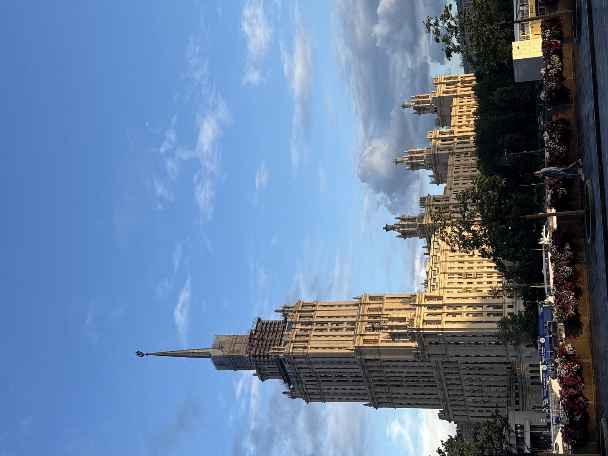 This image shows the Stalinist-era skyscraper, the Hotel Ukraina (now Radisson Collection Hotel, Moscow) in Moscow, Russia. The building is a prominent landmark of the city, characterized by its ornate architecture and spire topped with a star. The sky is blue with scattered clouds, and the building is bathed in the warm light of what appears to be either sunrise or sunset. In the foreground, a row of colorful flower arrangements is visible, bordering a walkway. Behind the flowers, a blue boat is docked on a body of water, possibly the Moskva River. A person is walking along the walkway. The surrounding area is green with trees, and other Stalinist-era buildings are visible in the background. The spire of the Hotel Ukraina is undergoing some scaffolding work.