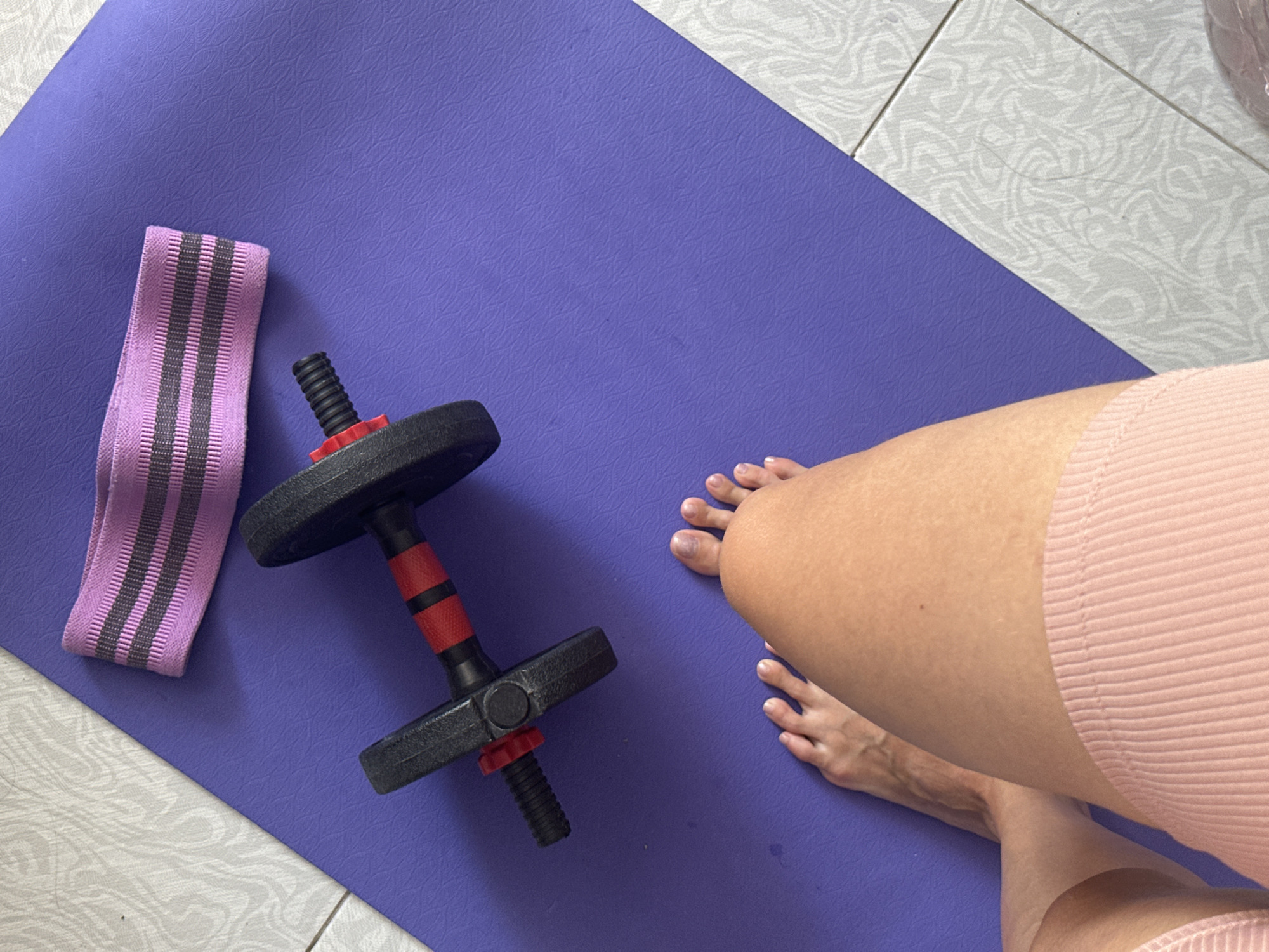 A pair of feet and part of a person's leg, wearing pink ribbed shorts, are positioned at the bottom left of a purple yoga mat. To the left of the mat, on a tiled floor, are the person's bare feet. On the yoga mat, a dumbbell with black and red accents is placed horizontally. Above the dumbbell, a pink resistance band with black stripes lies horizontally. The scene is indoors, with the tiled floor suggesting it might be a home or gym environment. No specific location in Sochi, Russia can be identified from the image.