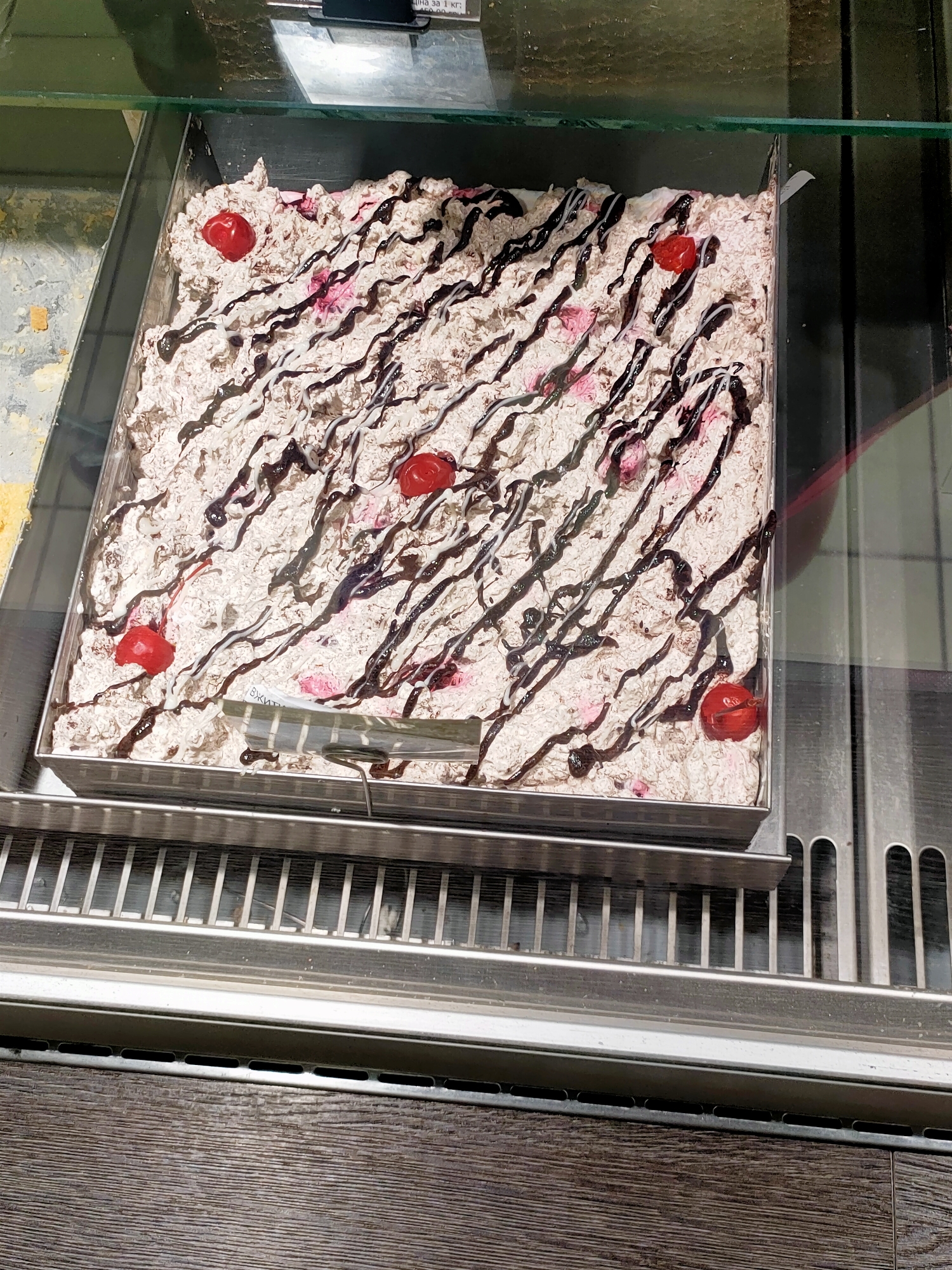 A square metal pan filled with a creamy dessert, possibly ice cream or cake, is displayed in a refrigerated glass case. The dessert is topped with a drizzle of dark chocolate sauce and appears to have pieces of fruit or cherries mixed in. Several whole cherries are placed on top as decoration. A small white label with some text is partially visible on the right side of the pan. The display case is situated in what appears to be a food establishment, indicated by the metal grating below and the wooden textured surface at the bottom of the image. The location is identified as Kyiv, Ukraine.