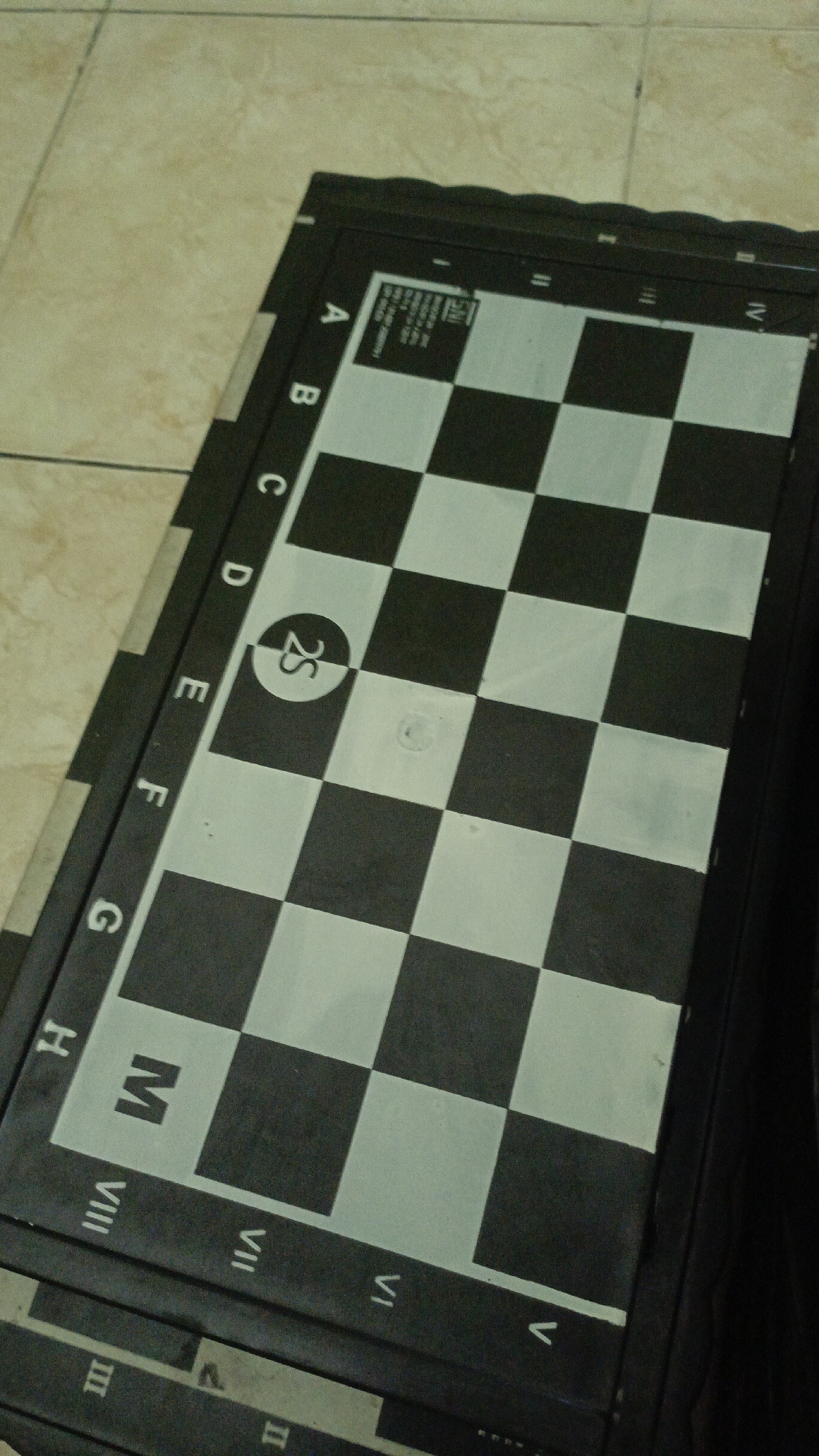 This is a close-up, high-angle shot of a black and white chessboard placed on a light-colored tiled floor. The chessboard is oriented diagonally, with the letters A through H visible along the left edge and Roman numerals I through VIII visible along the bottom edge. Some of the squares are a light gray, while others are black. There is a circular emblem with the characters "2S" in white within a black circle on one of the light gray squares. The chessboard appears to be made of plastic or a similar material and is slightly curved, possibly indicating it is a roll-up board. The floor tiles are rectangular and laid in a staggered pattern. The overall lighting is dim, with some glare on the surface of the chessboard. There are no people or specific locations in Trihanggo, Indonesia, visible in the image.