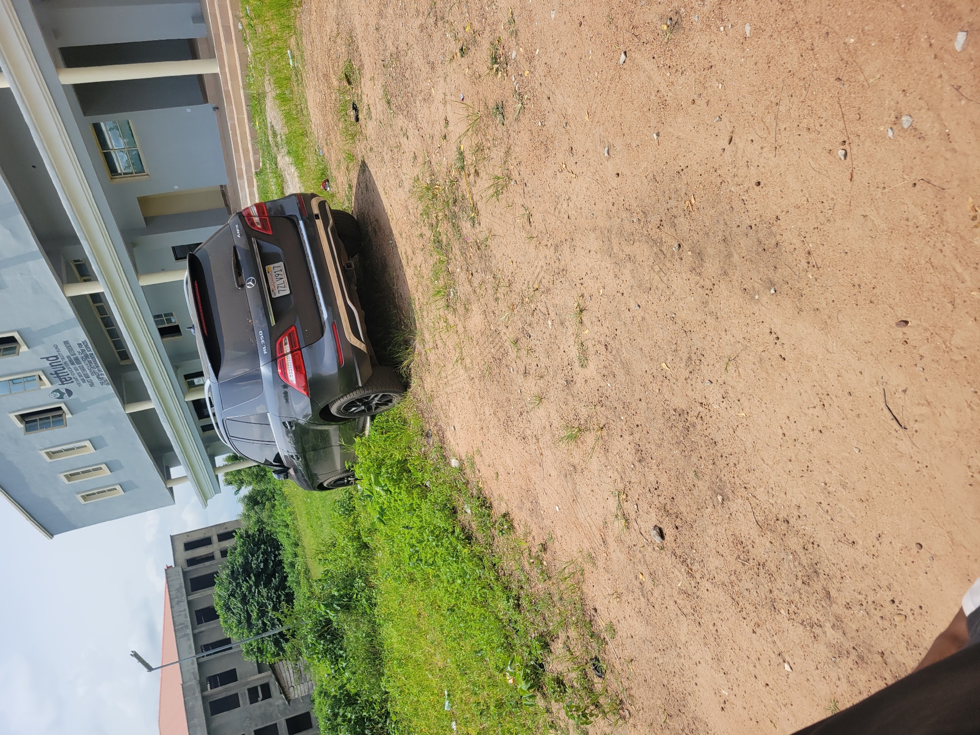 In an outdoor scene, a dark gray Mercedes-Benz ML 350 SUV is parked on a dirt and grass lot. The vehicle, with license plate "7ZL V917", is positioned facing slightly away from the viewer, with its rear visible. Behind the car, a light blue two-story building with a white trim and columns stands prominently. The building has multiple windows and an entrance area with stairs. To the left of the blue building, another partially constructed gray building with visible metal framework and red roofing is present. The environment appears somewhat unkempt, with overgrown green grass and scattered dirt. The sky is overcast. The image seems to have been taken during the daytime.