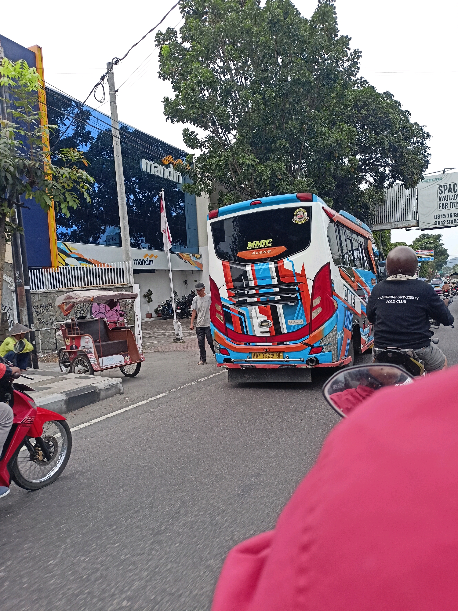 In the city of Magelang, Indonesia, a bus with a vibrant orange, blue, and white design is parked on the right side of a road. The bus has "MMTC Avenda" printed on its rear. A man in a gray t-shirt and dark pants stands beside the bus. To the left of the bus, a traditional three-wheeled passenger vehicle, known as a "becak," is parked. Two individuals are seated in the becak, and another person wearing a conical hat is crouched nearby. A red motorcycle is visible in the foreground on the left, with its wheel facing the viewer. In the background, a modern building with a blue glass facade displays the "mandiri" logo. Trees and a billboard with advertising are also present. The scene appears to be a street in an urban area, possibly near a commercial district.