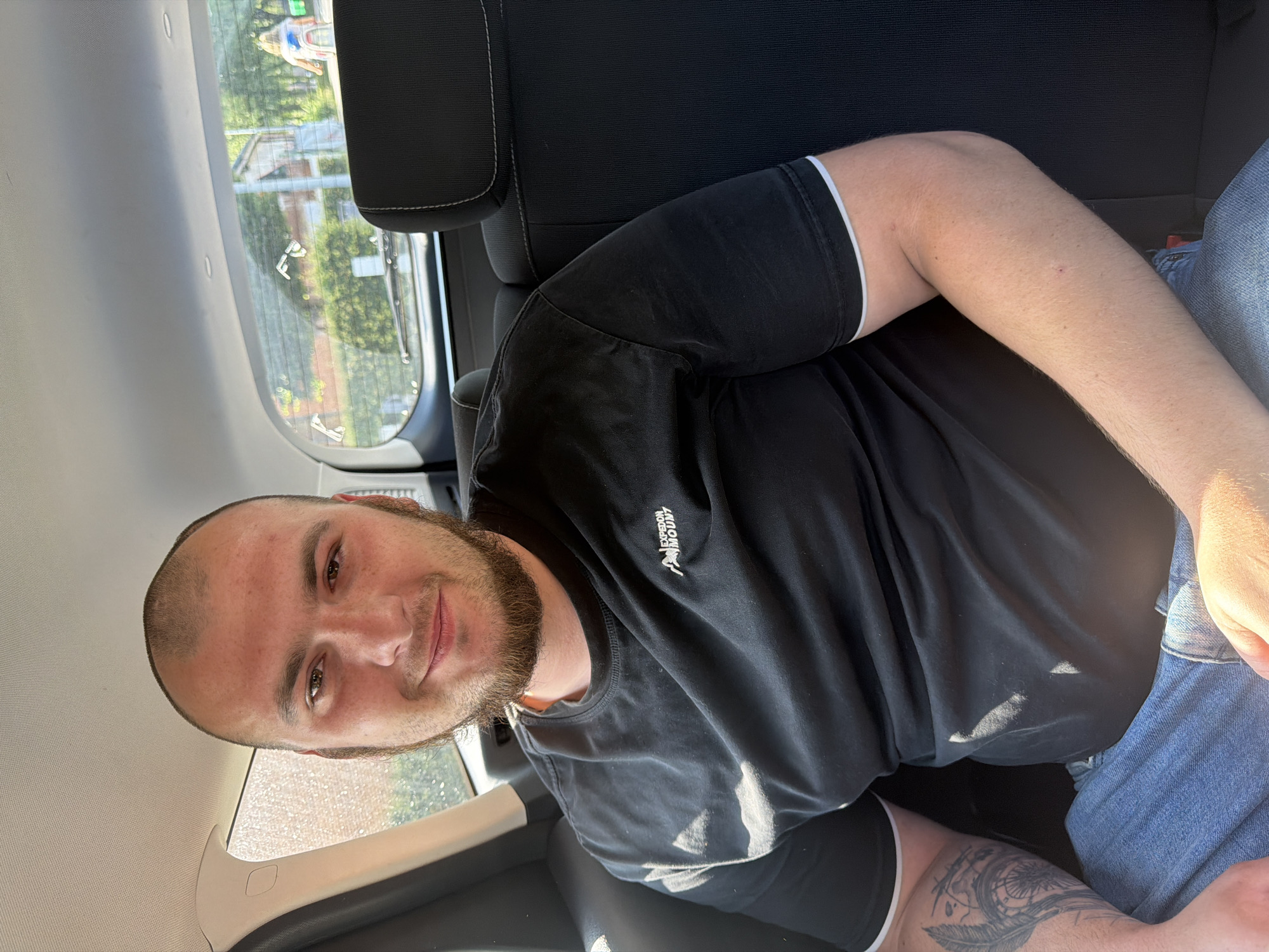 A bald, bearded man with light brown eyes sits in the backseat of a car, smiling at the camera. He is wearing a black t-shirt with a white trim on the sleeves and blue jeans. A tattoo of a feather and a dreamcatcher is visible on his left arm. The interior of the car is visible, including black seats and a sun-drenched window. Outside, a residential area with trees and a street can be seen. The scene appears to be located on a sunny day in Ravels, Belgium.