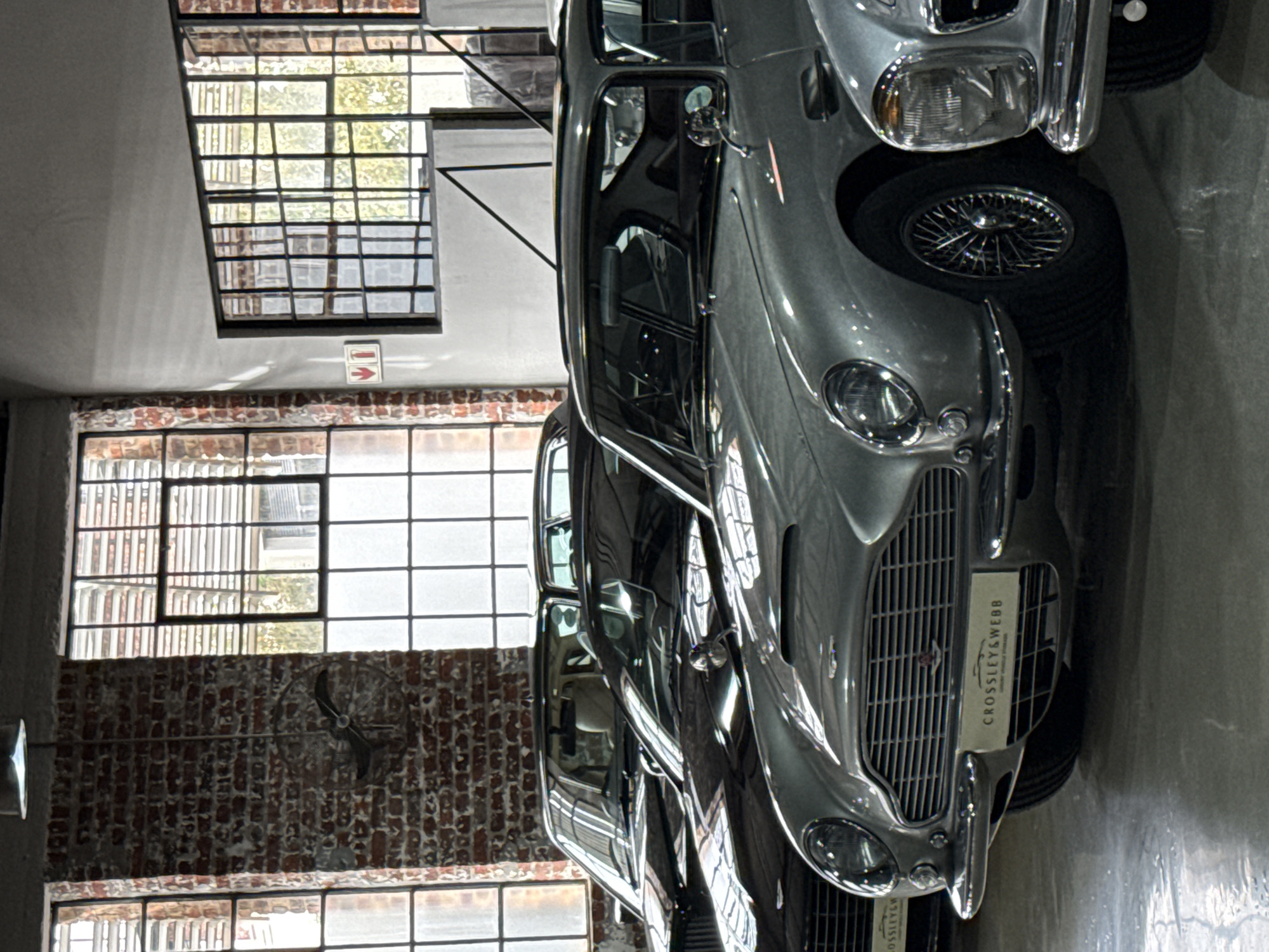 The image displays the interior of a luxury classic car showroom or dealership, likely in Cape Town, South Africa, characterized by an industrial-chic aesthetic. The main subjects are several vintage automobiles, lined up and on display.
In the foreground, a prominent silver-grey classic car, possibly an Aston Martin due to its iconic grille and styling, faces towards the left of the frame. It features gleaming chrome bumpers, a large grille, and visible headlights. A white rectangular plate holder on its grille clearly reads "CROSSLEY & WEBB" in black uppercase letters, with "LUXURY SPORTS & CLASSIC" printed in smaller text below it. The car's front right wire wheel is partially visible. Behind this vehicle, another similar classic car is parked, and a third car is partially visible further back, suggesting a collection.
The setting features large, multi-pane industrial-style windows spanning the left wall, allowing significant natural light into the space, indicating it is daytime. The windows offer glimpses of greenery outside and reflect the interior. Portions of the interior walls are made of exposed red brick, particularly below the large windows, contributing to the industrial feel. A vintage-style ceiling fan is mounted on one of these brick walls. Above a lower section of the windows, on a white wall, a small white sign with a red arrow pointing right is visible, likely an indicator or directional sign. The floor appears to be concrete.
No people are present, and the cars are stationary, suggesting they are being showcased or stored. The scene evokes a sense of elegance, nostalgia, and appreciation for classic automotive design and heritage.