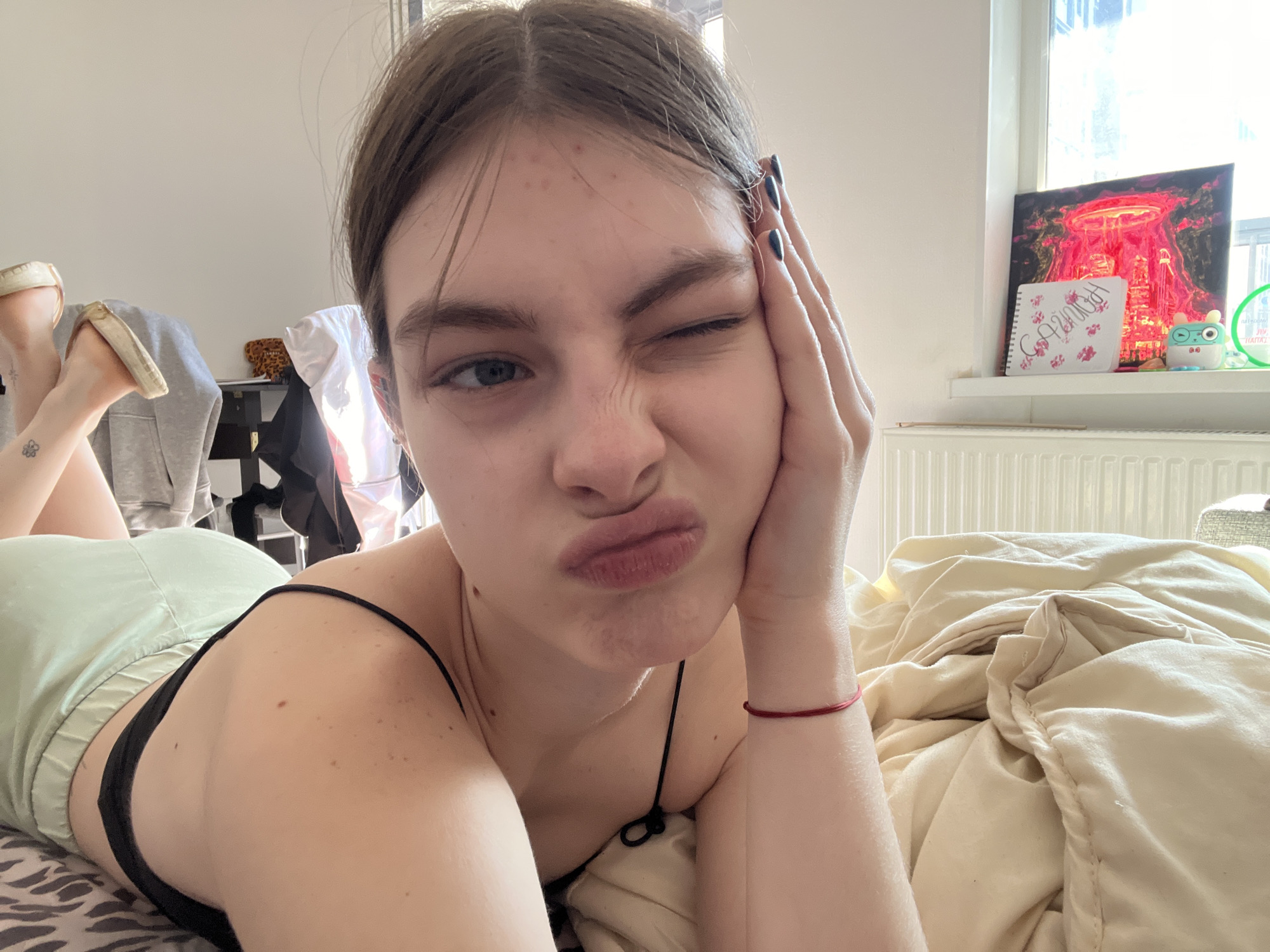 A young woman lies on a bed with patterned sheets and a light-colored duvet, wearing light green shorts and a black spaghetti strap top. She is winking her left eye and pouting her lips while resting her face on her right hand, which has dark nail polish. In the background, a window is visible with a cityscape, and on the windowsill, there is a painting of a city skyline with a volcano-like eruption, a notebook with writing, and a small toy. In the far back left, a pair of tan slip-on shoes and a grey hoodie are visible. There is no verifiable information to confirm the location as Saint Petersburg, Russia.