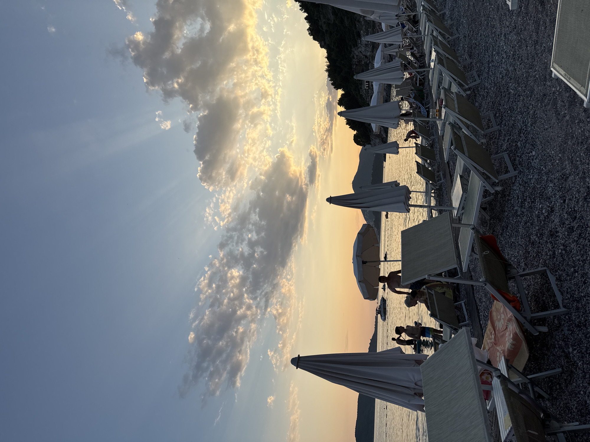 This media file depicts a beach scene at Kamenovo, Montenegro, during what appears to be late afternoon or early evening. The sky is a prominent feature, with a dramatic display of clouds illuminated by the setting sun, casting golden hues and creating visible sunbeams that radiate across the sky.
The foreground is occupied by a pebble beach lined with numerous white beach umbrellas and lounge chairs, many of which are empty. The sea is calm and reflects the warm light of the sky. Several people are visible interacting with the water and the beach. One person is partially submerged in the water, appearing to bend down. Another group of people, including adults and children, are standing near the water's edge. Some are silhouetted against the bright background.
The overall atmosphere suggests a peaceful end to a day at the beach, with the warm lighting contributing to a serene and relaxing mood. The lush green hillside forms the backdrop to the beach, further grounding the scene in a natural, scenic environment. There is no visible text in the image.