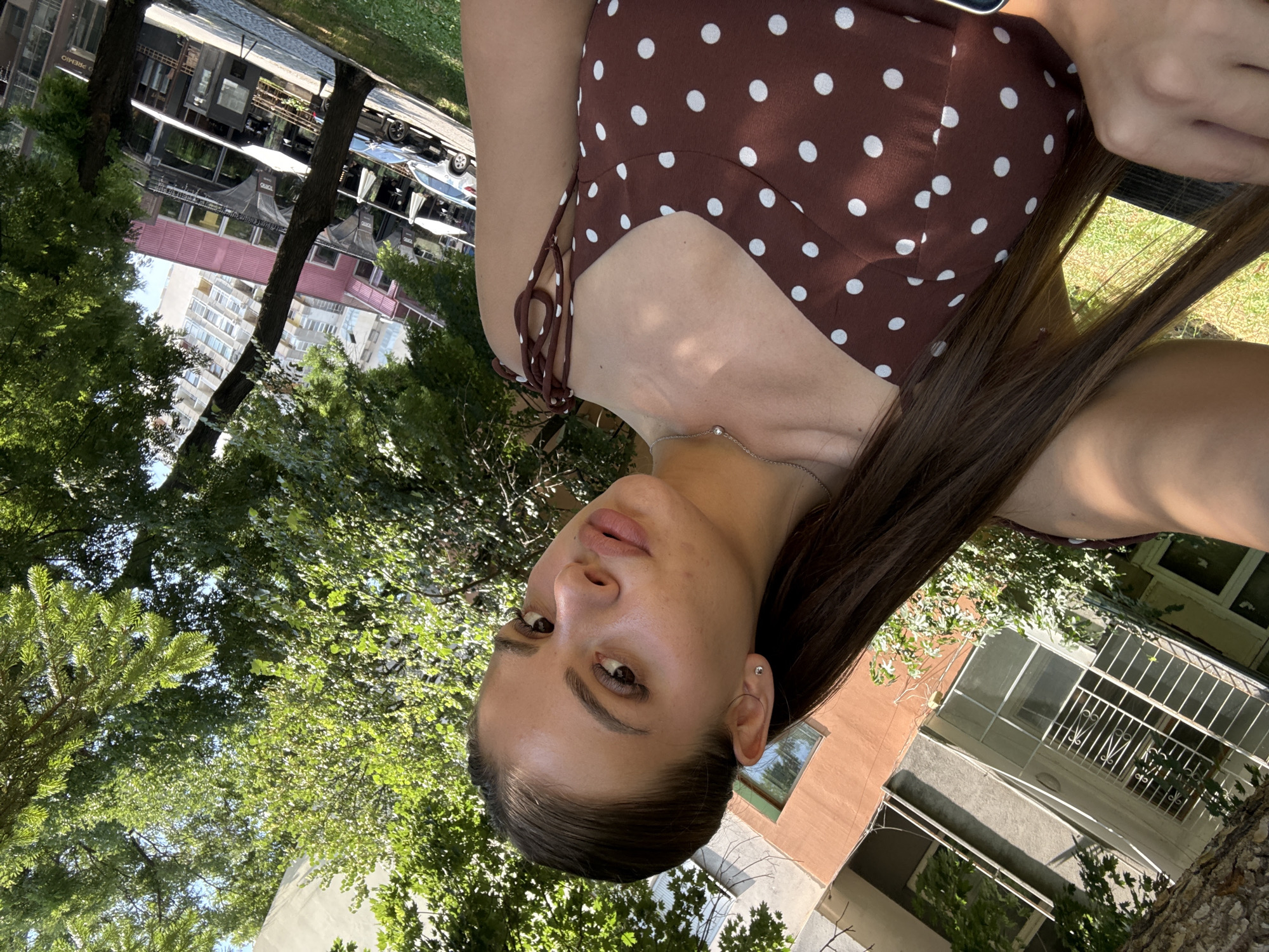 A young woman with long brown hair pulled back in a ponytail, wearing a brown polka-dot dress, takes a selfie outdoors. She has a delicate necklace on and a small stud earring. In the background, trees with green leaves are visible, along with buildings. One building appears to be a residential apartment complex with balconies and windows, while another has a pink facade and a black awning over storefronts. Cars are parked on a paved area next to the building with awnings. The scene suggests an urban environment, possibly a street or a park area within the city of Plovdiv, Bulgaria, given the architectural style and vegetation. The lighting indicates it is daytime.