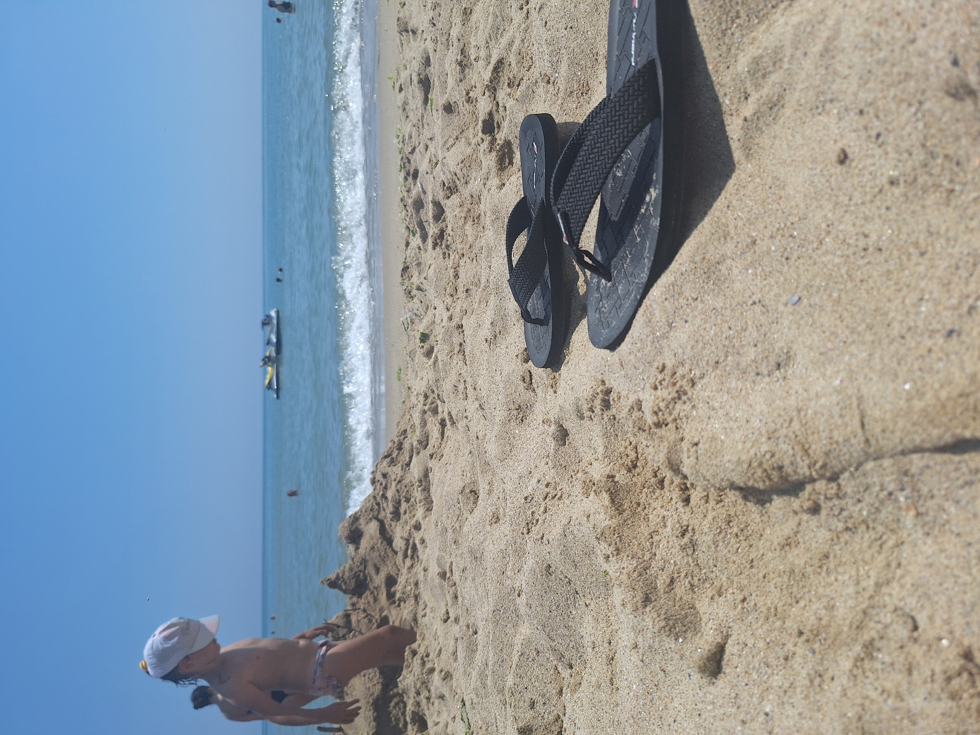 The image depicts a sunny day at Bliznatsi Beach, Bulgaria. In the foreground, a pair of black flip-flops rests on the sand. Behind them, a child wearing a white cap and patterned swim trunks is seen playing with a sandcastle. Further in the distance, the calm blue sea stretches to the horizon, with a few jet skis and people visible in the water. The overall scene conveys a typical summer beach atmosphere.