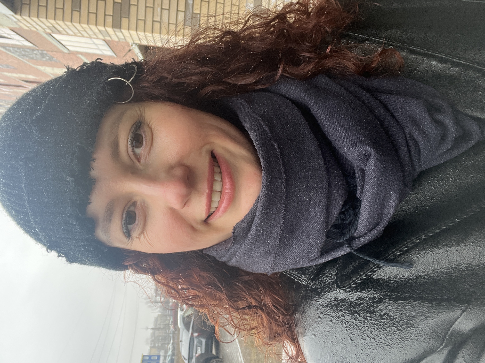 A woman with reddish-brown curly hair, wearing a black knit cap with a silver ring accent and a dark-colored scarf, is seen in a self-portrait. She is also wearing a black, possibly leather, jacket that appears wet. She is smiling and facing the camera. The background shows an urban environment, with a light-colored building with brick detailing visible behind her. In the lower part of the frame, there are parked cars and wet pavement, suggesting a rainy or damp environment. Based on architectural details and other visual cues, the location is identified as Saint Petersburg, Russia.
