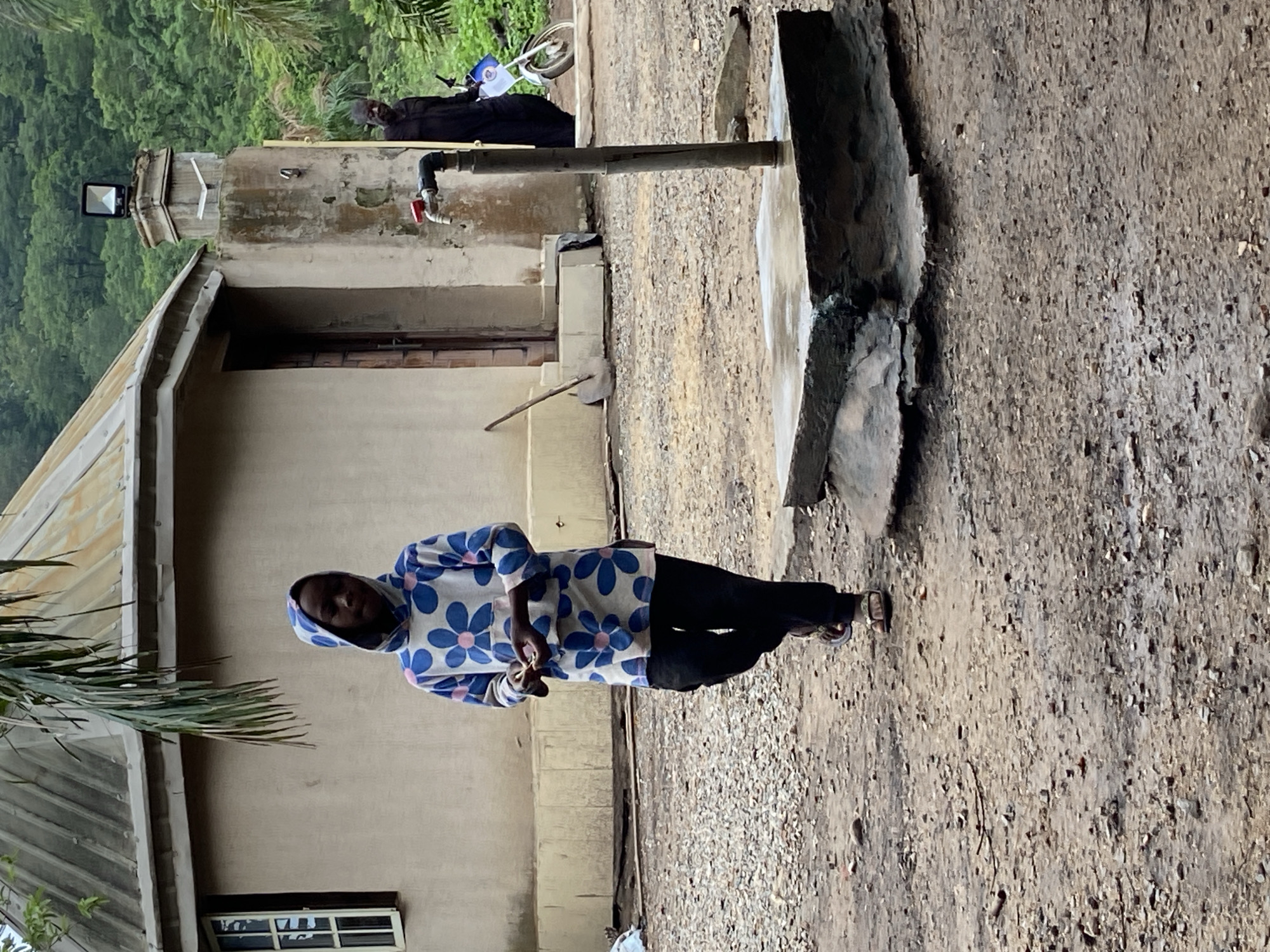 The image captures an outdoor scene in Mararaba, Nigeria, featuring two individuals and a residential structure.
In the foreground, slightly left of center, a young person with dark skin walks towards the right. They are dressed in a white hooded long-sleeved top adorned with a repeating pattern of blue and pink flowers, dark trousers, and light-colored sandals. Their gaze is directed downwards, focused on a small, indistinct object being manipulated between their hands.
To the right of the young person, a functional water tap structure stands. It comprises a vertical metal pipe extending from a rectangular concrete base, which shows weathering and dampness. A red-handled faucet is positioned horizontally on the upper section of the pipe.
In the mid-ground, a single-story building with light-colored, possibly stuccoed, walls is visible on the left. The structure has a distinct rounded or polygonal form with a sloping, corrugated metal roof exhibiting signs of age and discoloration. A white-framed multi-pane window is located on the left side of the building, and a dark wooden door is visible to the right, accessible by a low, light-colored step. A long-handled shovel leans against the building wall near the door.
In the background, on the right side, an adult male with dark skin and a beard is partially obscured by a weathered concrete pillar. He wears a dark, long-sleeved garment. The pillar supports a black rectangular floodlight on its apex and has a small white, angular object attached to its side. A white motorcycle, partially visible next to the man, features a small blue and white flag or banner attached to its rear.
The ground across the scene is unpaved, composed of packed dirt and scattered gravel and small stones, with some areas appearing damp. Lush green foliage, including a large palm frond hanging from the top of the frame and dense trees covering distant hills, forms the backdrop, indicating a heavily vegetated environment.