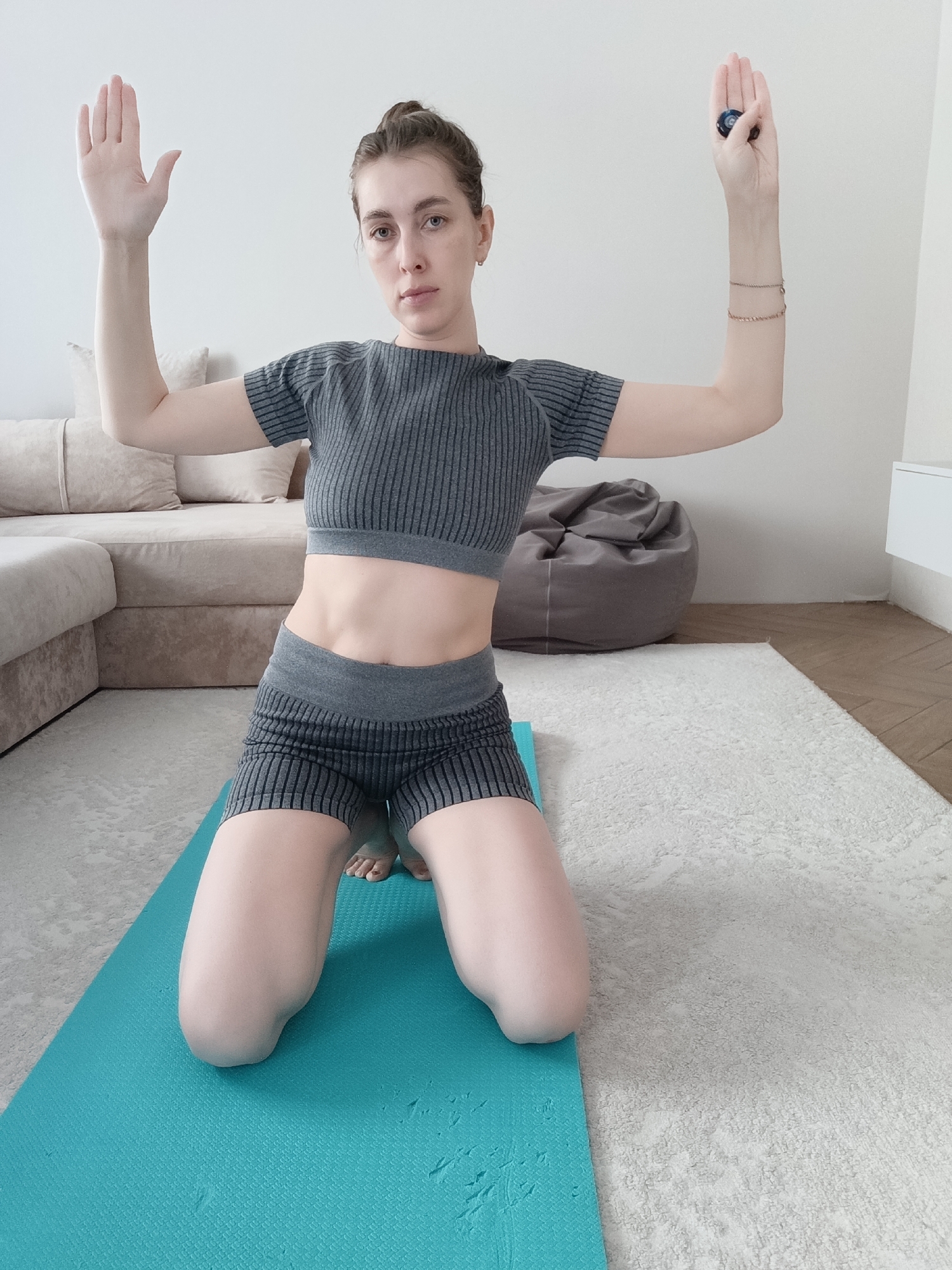A woman kneels on a teal yoga mat in an indoor setting. She is wearing a matching ribbed gray crop top and shorts set, with her abdomen visible. Her hair is pulled back, and she gazes directly at the viewer. Both arms are raised; the left hand is open and facing forward while the right hand holds a dark blue object. Two thin gold bracelets adorn her right wrist. Behind her, there is a neutral-toned sofa, a gray beanbag chair, a white cabinet, and a white wall. The floor appears to be wood in the corner near the cabinet and covered by a patterned white rug under the yoga mat and sofa. The setting suggests a room in a home or apartment, located in the city of Chaikovsky, Russia.