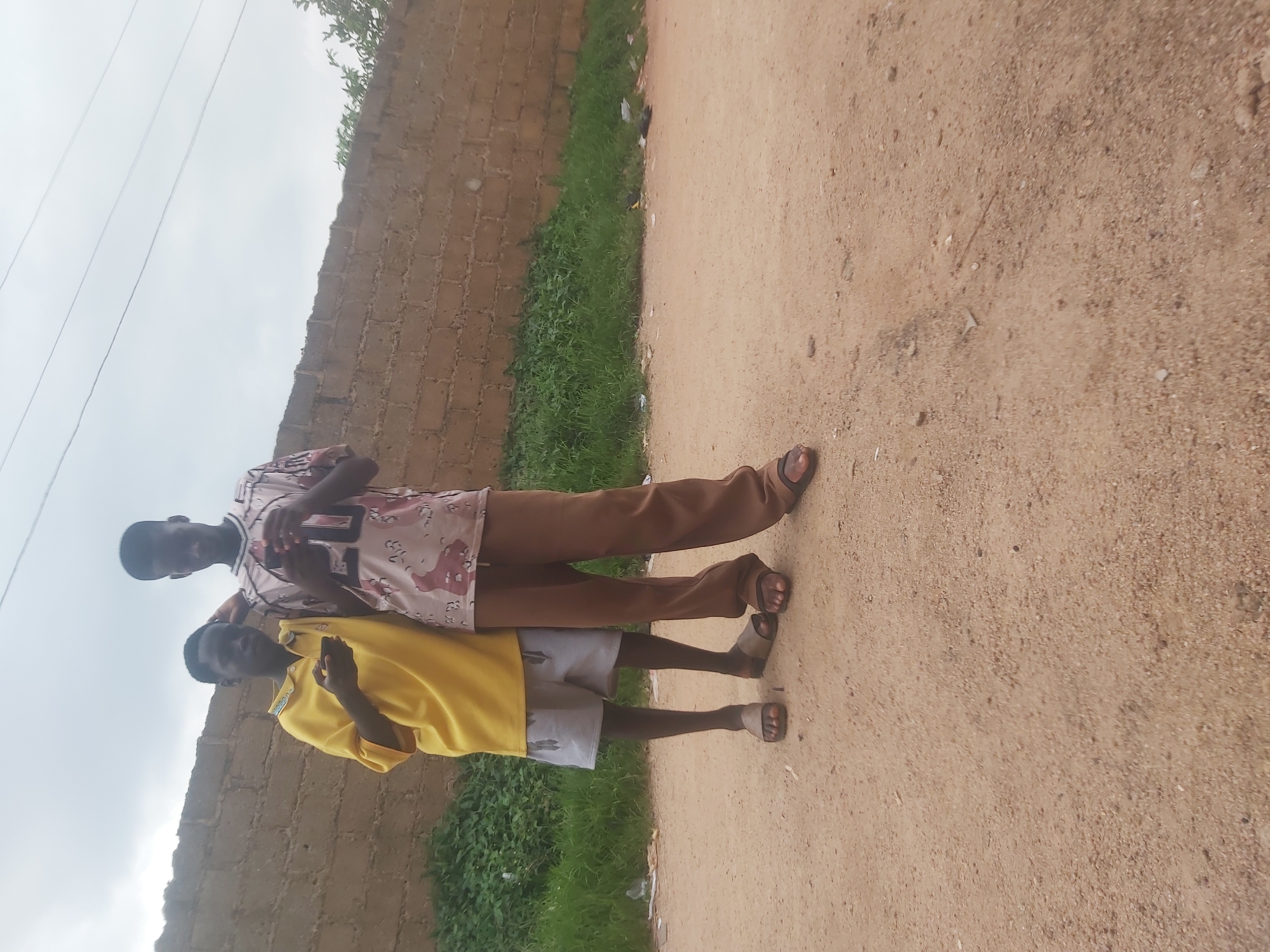 Two young males stand outdoors in front of a brick wall. The male on the left wears a bright yellow t-shirt and grey shorts. He is looking down at his phone, which he holds in his left hand. The male on the right wears a patterned t-shirt and brown pants. He is also looking down at his phone, which he holds in his left hand. Both males are wearing sandals. The ground is a dirt path, and there is green vegetation behind the brick wall. The sky is overcast. The location is believed to be in Jos, Nigeria.