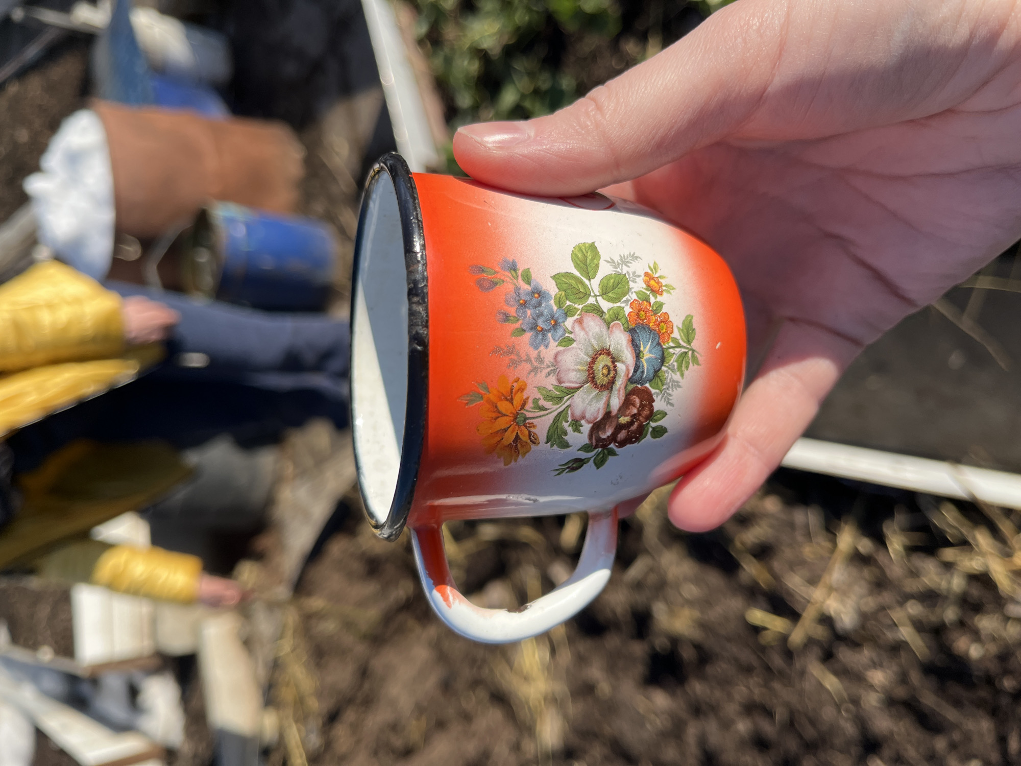 A hand holds an enameled metal mug with a floral design. The mug has a white background that fades into orange near the rim and base. The floral design includes flowers of various colors: blue, orange, white, and brown. The mug’s rim is black, and the handle is white. In the background, out of focus, are objects scattered on the ground, including what appears to be clothing in shades of yellow and blue, and debris. The ground itself is brown and looks like dirt or soil. The scene is illuminated by natural light. There is no information to indicate the location is the city of Chaikovsky, Russia.