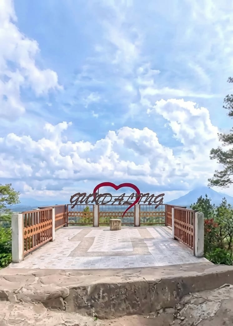 The image shows a scenic viewpoint at Gundaling 2, Indonesia, featuring a raised platform with a decorative railing and a large sign with a heart shape and the text "Gundaling". The platform is paved with tiles and has a rustic stone border in the foreground. In the background, rolling hills and a conical mountain are visible under a sky filled with fluffy white clouds. The platform appears to be empty of people.