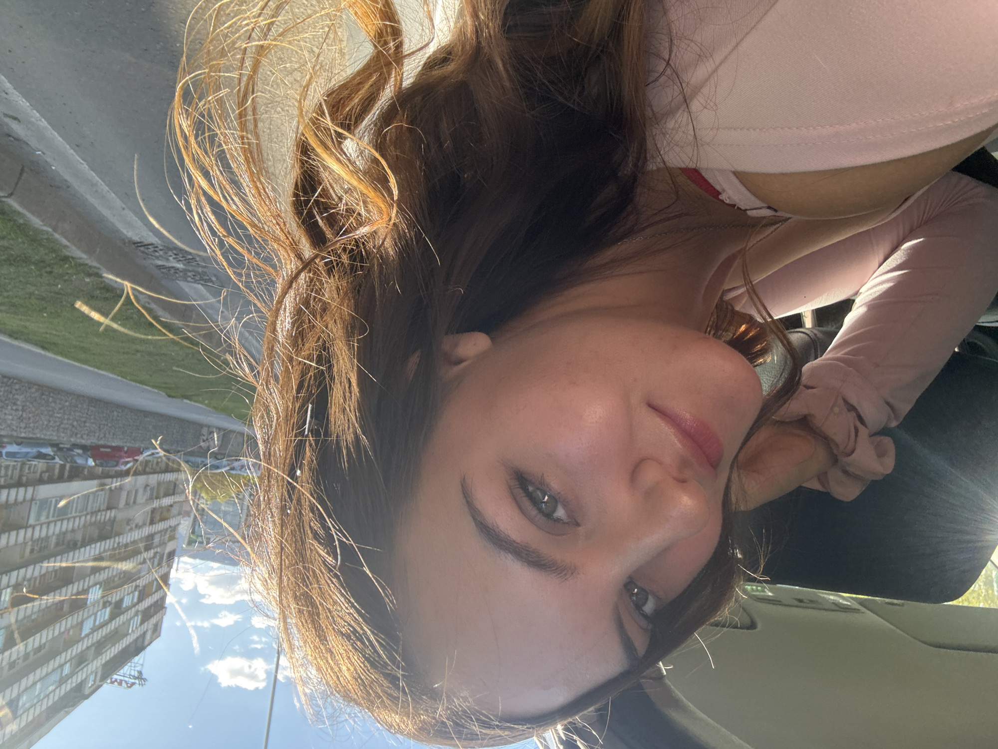 The image features a young woman inside a car, looking towards the camera with a gentle, slightly playful smile. Her head is tilted, suggesting a casual, low-angle selfie or portrait. She has long, wavy brown hair with reddish-brown highlights, strongly illuminated by bright natural sunlight that creates a soft lens flare on the right side and highlights individual hair strands. Her skin is fair with light freckles, and she has expressive green or hazel eyes. She is wearing a light pink or peach-colored long-sleeved top and a thin silver necklace.
The setting combines elements of a car interior and its reflections. Inside the car, parts of a dark surface, a grey headliner, and a car visor are visible, with a light pink scrunchie hanging from the visor. The car window reflects an urban or suburban environment. These reflections include multi-story residential buildings with numerous windows and balconies, a grey road or pavement bordered by green grass, and faint shapes of other vehicles. The sky reflected is bright blue with scattered white clouds, indicating a sunny daytime scene.
On one of the reflected buildings, faint text "AM" is discernable, likely part of a sign. The architectural style of the buildings and the general urban environment are consistent with the provided location context of Sofia, Bulgaria. The strong natural light and clear sky suggest the photo was taken during the day, likely morning or afternoon. The woman's expression conveys a sense of calm and slight contentment.