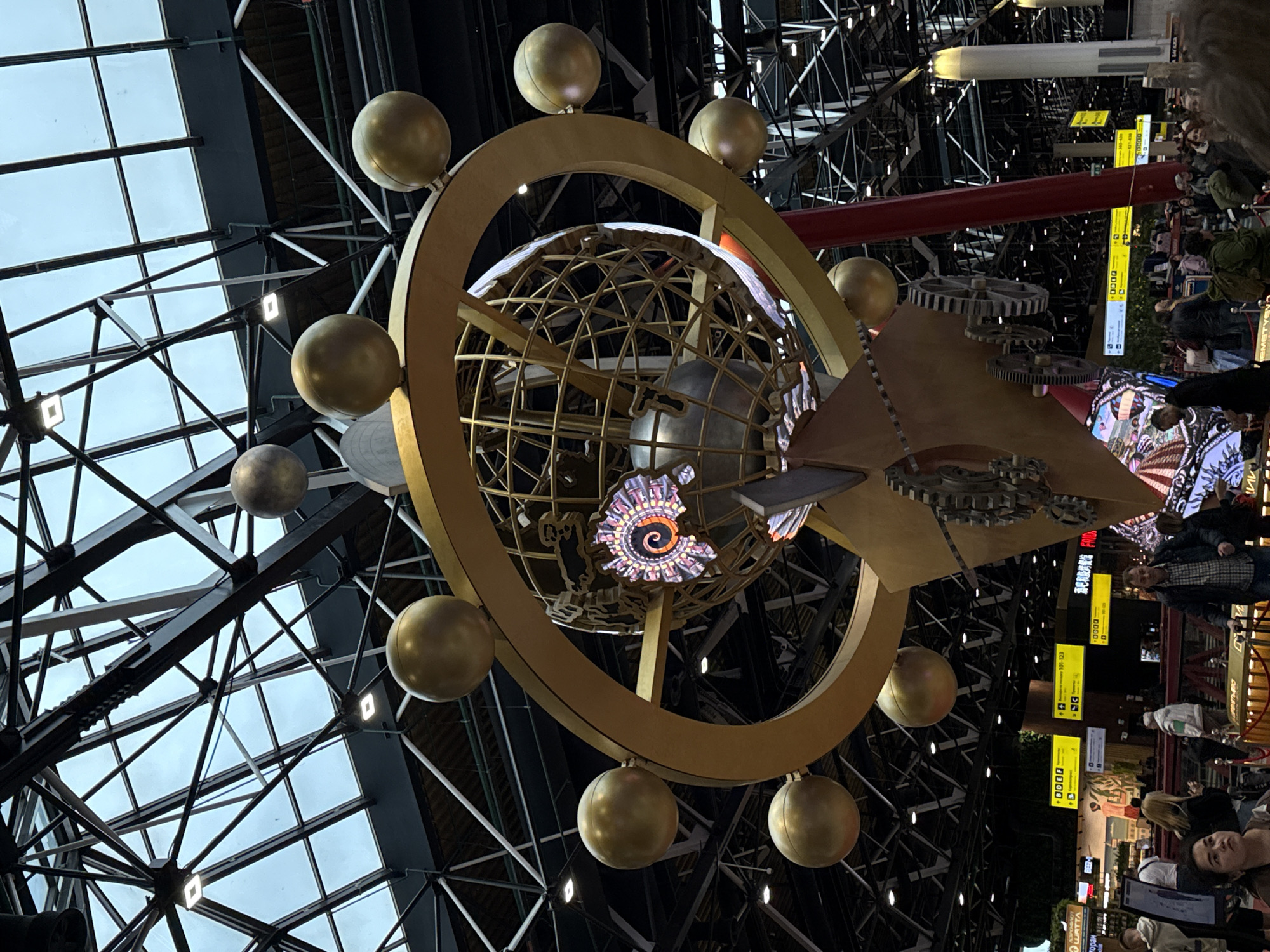This media file captures a large, ornate golden sculpture suspended within a vast indoor public space, likely Sheremetyevo International Airport in Khimki, Russia. The shot is taken from a low angle and is slightly tilted to the right, looking up towards the ceiling.
The central subject is a multi-layered, armillary sphere-like sculpture, predominantly golden. It features a large outer ring adorned with numerous smaller golden orbs. Inside this ring, a cage-like golden globe is visible, with a luminous screen displaying a colorful, intricate spiral pattern at its core. On the right side of the sculpture, industrial-style gears and a pointed, arrow-like structure are integrated. Smaller, muted silver spheres are also part of the design, hanging slightly below the main golden structure.
The background reveals a towering, complex ceiling composed of dark metallic trusses and large glass panels, through which a bright, cloudy sky is visible, indicating daytime. Square-shaped artificial lights are embedded within the ceiling structure.
In the lower right and further background, numerous people are visible, appearing to be walking, standing, and waiting, suggesting a busy transit area. They are dressed in casual attire. A long, red cylindrical structure and a white cylindrical object are also seen extending across the upper background.
Visible text includes several bright yellow directional signs with black lettering. One sign partially shows "0000" with an arrow. Another displays "101-123" with an arrow, likely indicating gate numbers. A fragment of a word resembling "C H A Y N A" is also discernible on one of the signs. The overall atmosphere is busy and functional, with the grand sculpture serving as a prominent artistic centerpiece.