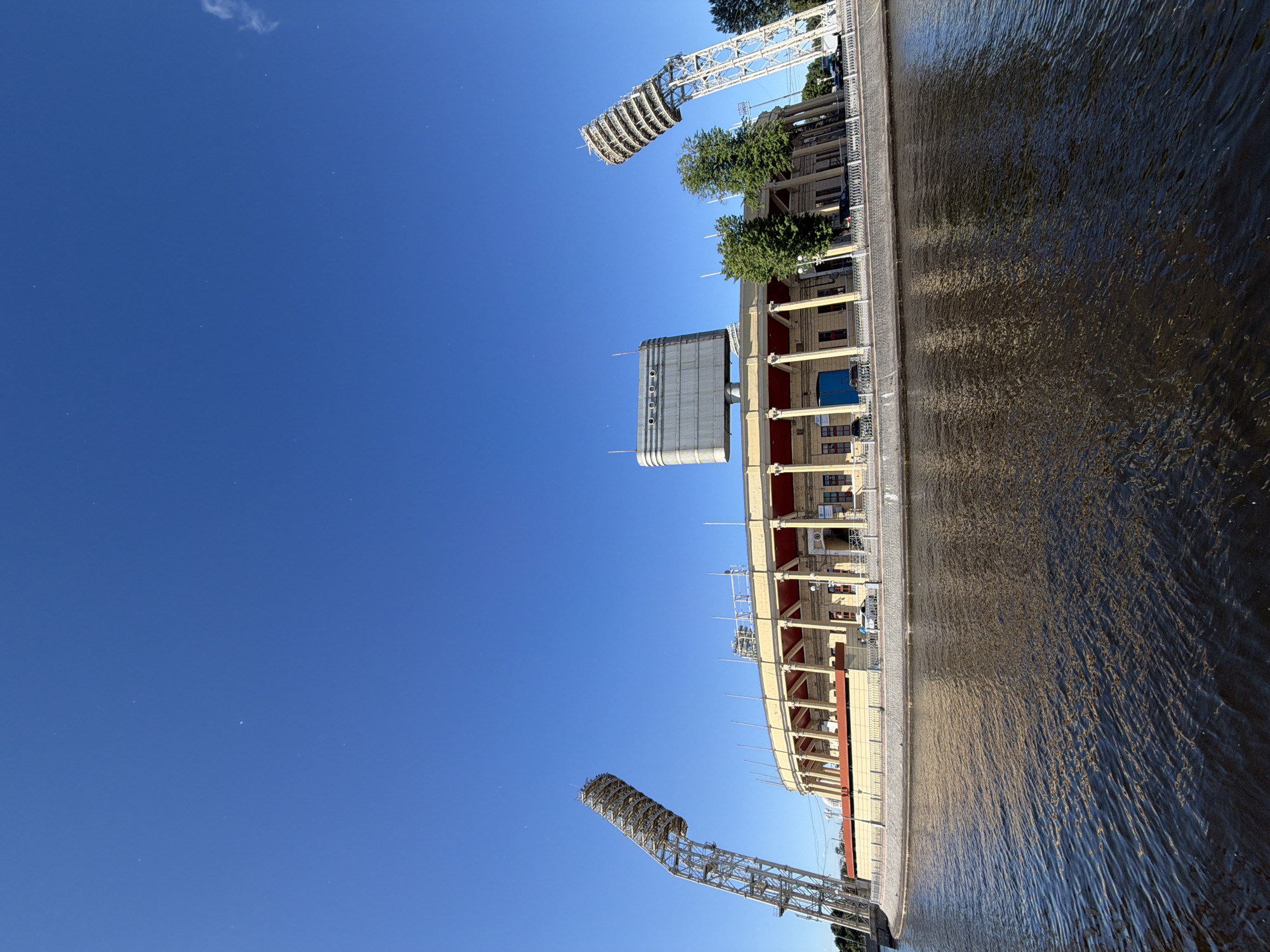 This is a wide-angle, low-angle shot of a stadium situated on the bank of a body of water, possibly a canal or river, in Saint Petersburg, Russia. The stadium, with its classic architecture featuring a colonnade and a distinctive large scoreboard, is illuminated by bright sunlight. Two tall, skeletal light towers flank the structure, one leaning inwards towards the stadium. The water in the foreground reflects the stadium's facade and the sky, which is a clear, deep blue with a few faint streaks of clouds. A low, decorative railing separates the stadium from the water's edge. A few vehicles and a couple of individuals are visible near the stadium, suggesting a public space. The overall impression is a serene, sunlit day at a prominent city landmark.