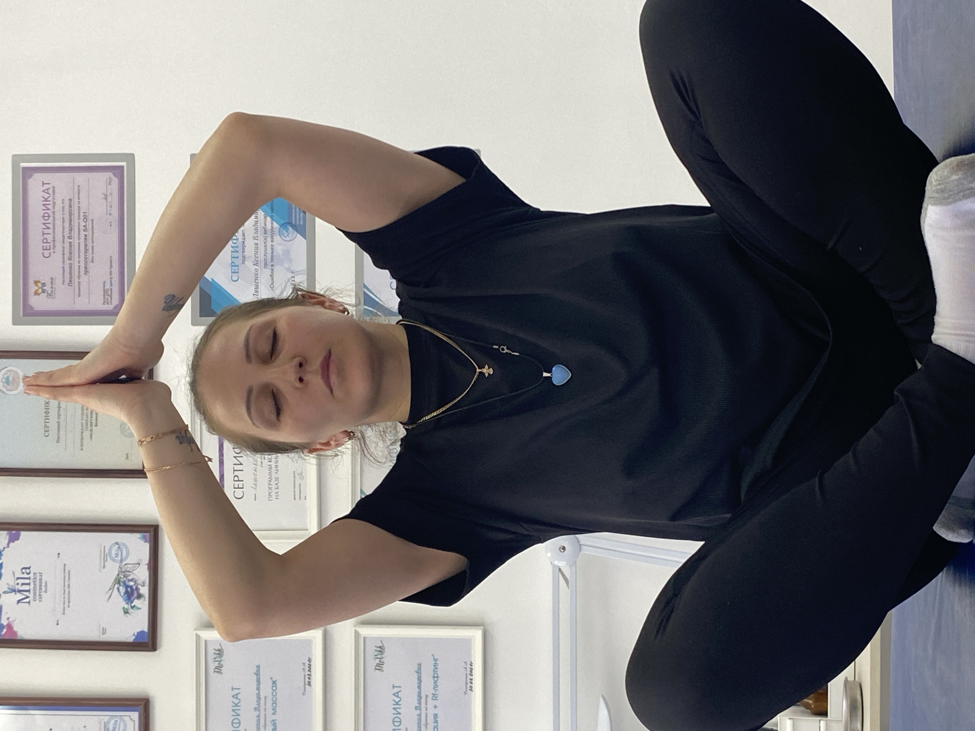 The image shows a woman wearing black leggings and a black t-shirt seated with her legs crossed and arms raised, her hands meeting above her head, possibly engaging in a yoga or meditation pose. She is also wearing a necklace. Behind her on the wall are several framed certificates and diplomas. Some text is discernible on them in the Russian language. Some of the titles read "CERTIFICATE" and contain Russian names such as "Ksenia Vladimirovna" and brands like "Mila cosmetics". One certificate mentions "RF-lifting". It's difficult to pinpoint the exact location in the city of Markovskiy, Russia based on the image.