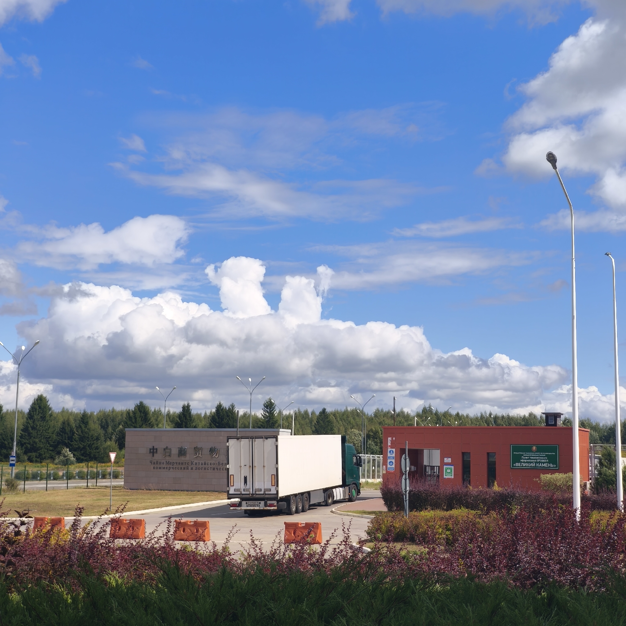 The image depicts a modern border crossing facility under a bright blue sky with scattered white clouds. A large, white, box-style truck with a green cab is positioned in the foreground, appearing to be in motion or recently stopped. To the left, a beige building with Chinese and Cyrillic lettering, identified as the "China-Belarus Commercial and Logistics Cooperation Zone" is visible. To the right, a red building with Cyrillic signage, which reads "Пункт таможенного оформления <<ВЕЛИКИЙ КАМЕНЬ>>" (Customs Clearance Point "Great Stone"), is present. The area is landscaped with low, red-leafed bushes in the foreground and a dense line of green trees forming a backdrop. Several tall, white streetlights are present, along with orange traffic barriers on the paved roadway. The scene suggests an active border checkpoint or logistics hub within the Драчкаўскі сельскі Савет in Belarus.