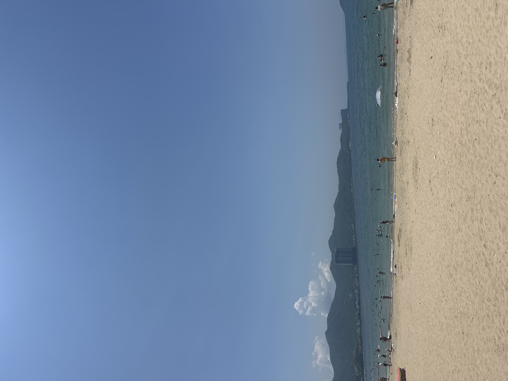 The image depicts a busy beach scene in Nha Trang, Vietnam, under a clear blue sky. The foreground is dominated by a wide expanse of golden sand. In the midground, a vibrant blue ocean stretches out, dotted with numerous people swimming, wading, and enjoying the water. Several individuals are visible on the beach, some standing, others sitting or lying down. A striped umbrella stands open near the water's edge, providing shade. In the background, lush green mountains form a scenic backdrop to the coastal city, with buildings and structures visible along the shoreline and nestled amongst the hills. The overall atmosphere is one of a lively summer day at a popular tourist destination.