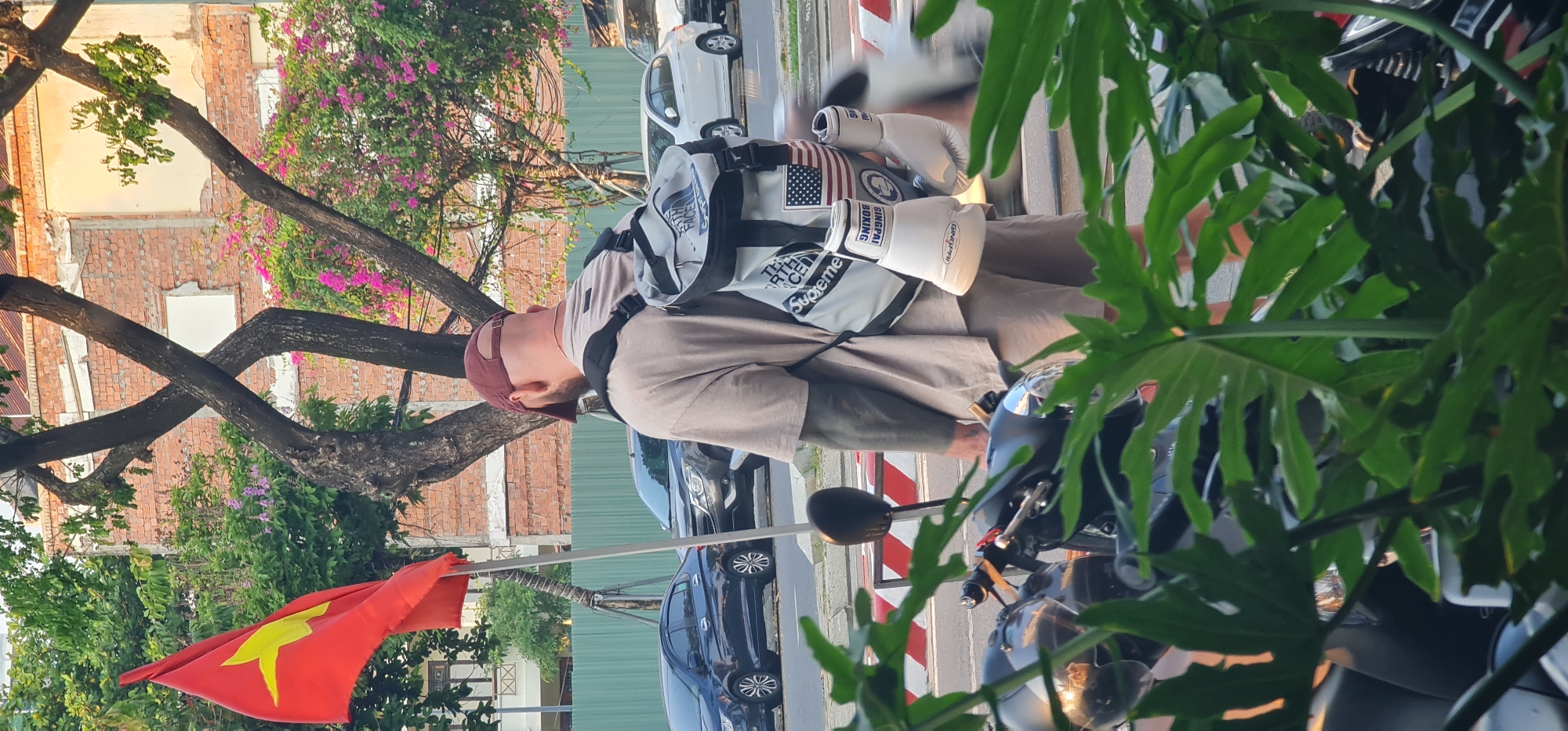 A man in a maroon baseball cap is standing in the city of Đà Nẵng, Vietnam. He is wearing a tan t-shirt, tan pants, and a backpack with a United States flag patch on it. He has a pair of white boxing gloves attached to his backpack. He is standing next to a black motorcycle and a Vietnamese flag is visible in the foreground. Buildings and trees are in the background. Several cars are parked on the side of the street.