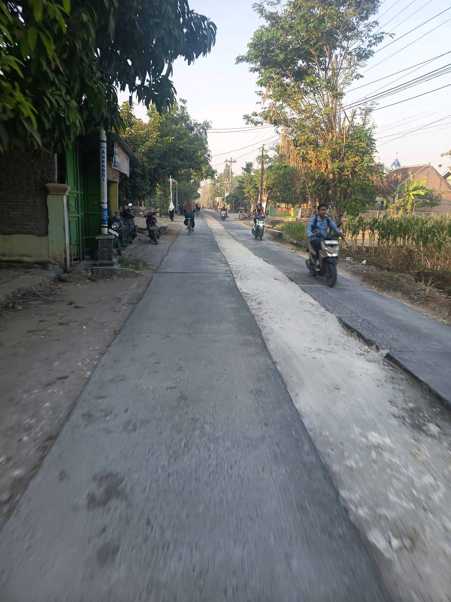 The image shows a road in Pulokulon, Indonesia, likely during daylight hours, with a clear sky. The road is paved, with a section of newer concrete adjacent to older asphalt, and shows signs of wear and repair. There are several people on the road. In the foreground, a young man in a blue shirt is riding a scooter. Further down the road, a woman in a hijab is riding a bicycle, and another person is riding a scooter. On the left side of the road, there are several parked motorcycles. To the left of the road is a building with green doors and a sign. Trees and vegetation are visible on both sides of the road, with power lines overhead. The scene appears to depict everyday street life in a residential or semi-rural area of Pulokulon.
