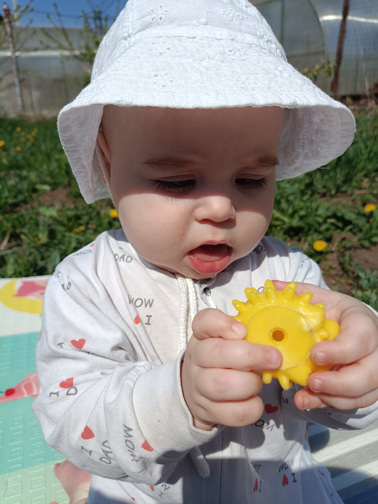 The image depicts a baby in Chaikovsky, Russia, wearing a white sun hat and a white pajama-like outfit with red heart and "I LOVE DAD" and "I LOVE MOM" prints. The baby is sitting outside, possibly on a playmat. The child is holding a yellow, sun-shaped toy with a hole in the center, looking down at it with a slightly open mouth. The background includes green grass with dandelions and a blurred fence and greenhouse. The lighting indicates a sunny day.