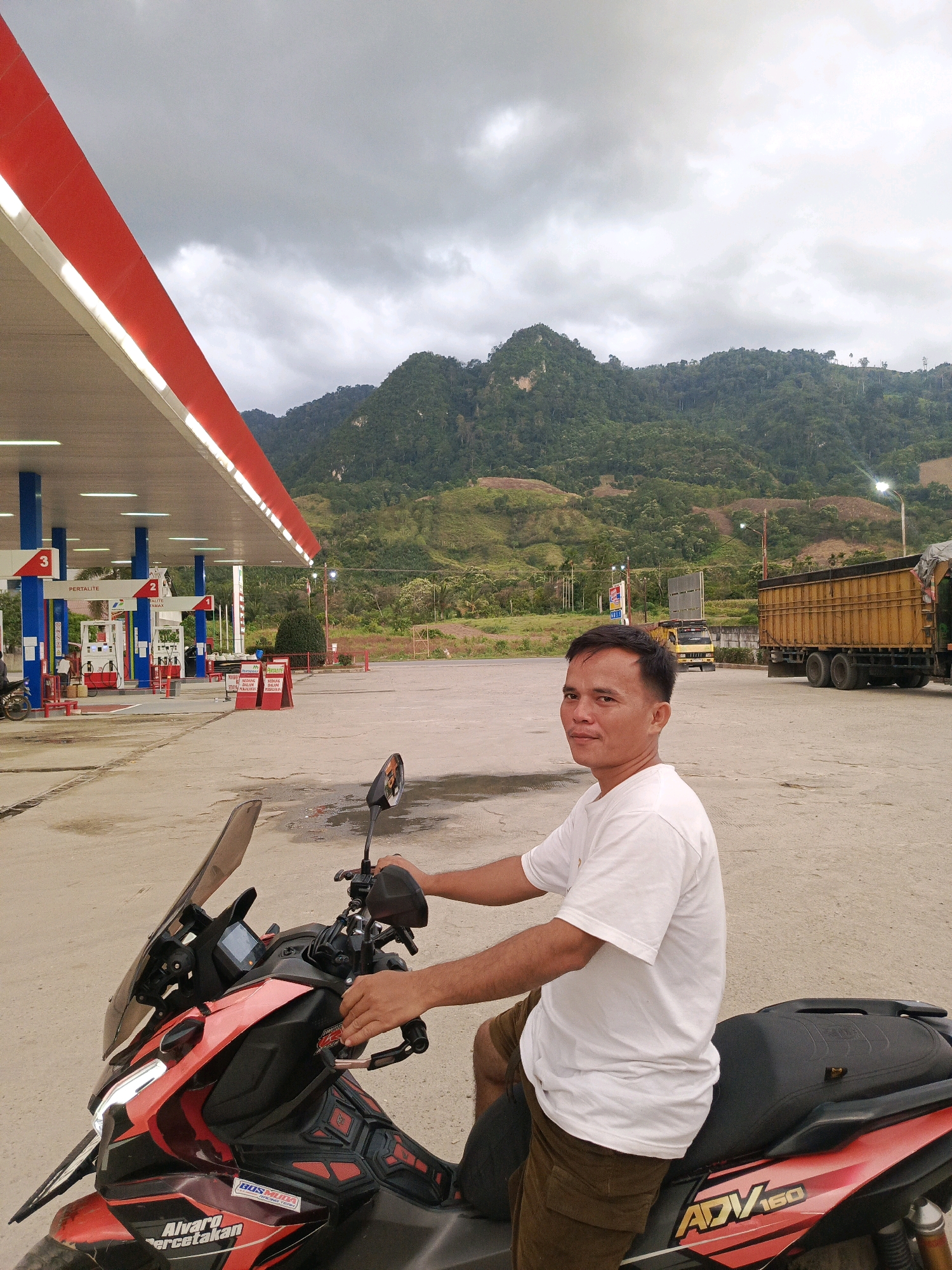 In Laubalang, Indonesia, a man sits on a red and black motorcycle at a gas station under a cloudy sky. The gas station features a red and white canopy supported by blue pillars, with gas pumps visible. In the background, a yellow truck and a larger yellow truck with a covered cargo bed are parked on the asphalt lot. Rolling, tree-covered hills dominate the distant landscape. The man, wearing a white t-shirt and brown shorts, is looking towards the camera. His hands are on the motorcycle's handlebars. The motorcycle has a windshield and various decals, including "ADV 160." The ground is damp in places, suggesting recent rain.
