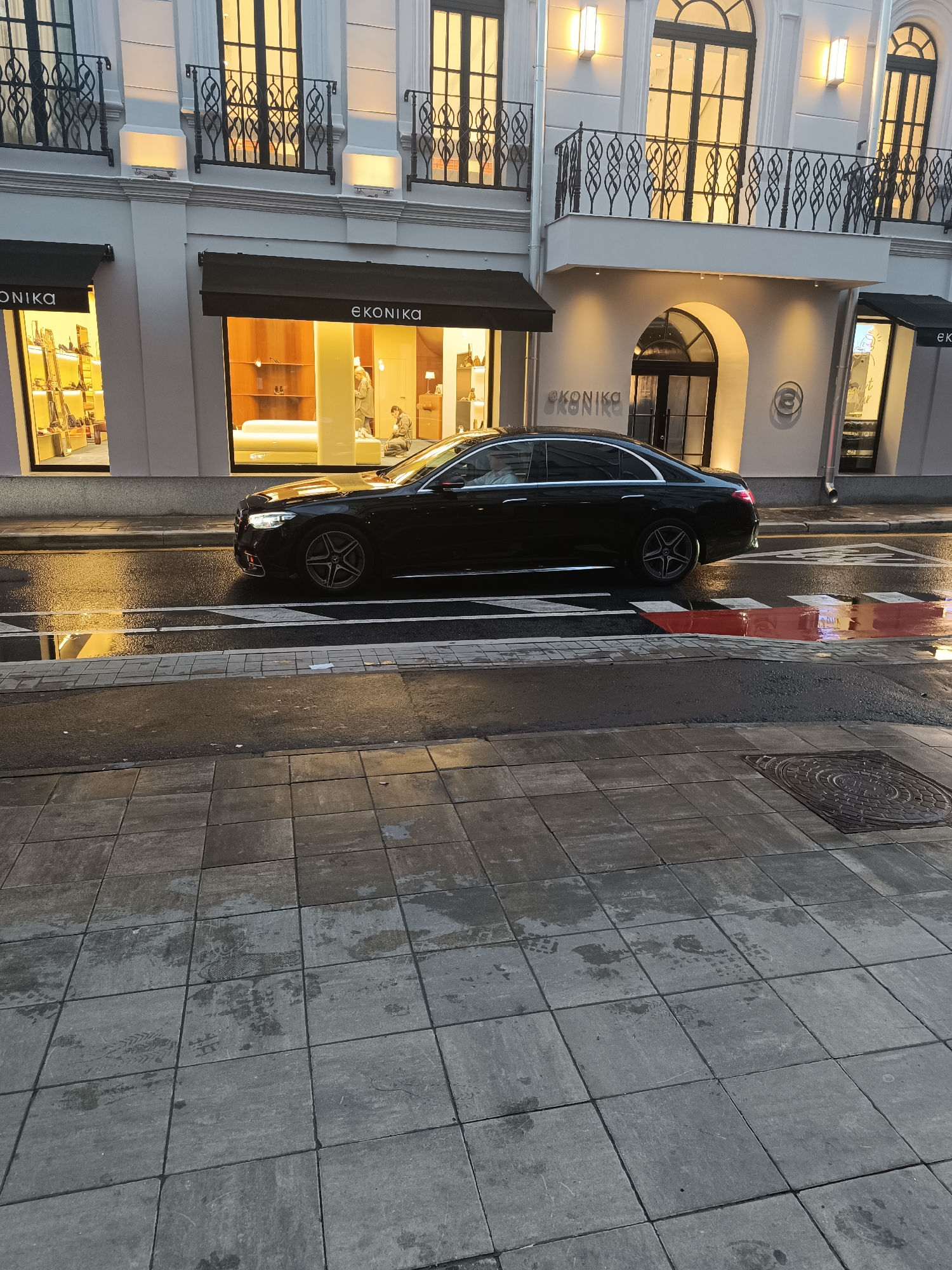 A black Mercedes-Benz S-Class sedan is parked on a wet street in front of a storefront with the name "EKONIKA" visible. The car's headlights are on, and it is facing left. The storefront has large windows displaying merchandise, and there are people inside the store. The street is paved with asphalt and has a pedestrian crossing with red markings. The sidewalk is made of gray square tiles, and there is a manhole cover visible on the sidewalk. The building behind the storefront has a white facade with large windows and decorative railings. The lighting suggests it is either early morning or late afternoon, with warm light coming from the windows and streetlights. The overall scene depicts a commercial street in a city, possibly after rain due to the wet conditions.