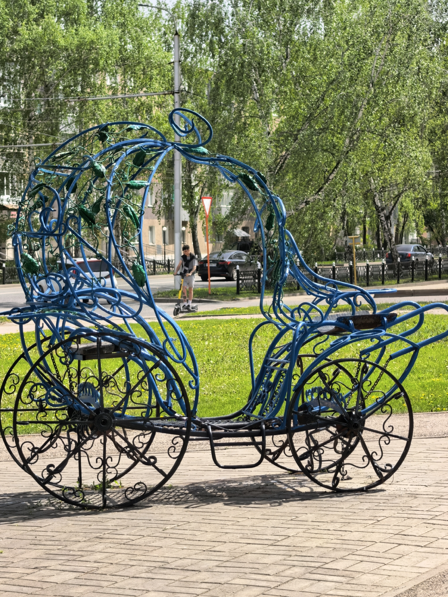 An ornate, blue, metal sculpture of Cinderella's carriage sits on a brick walkway in Sterlitamak, Russia. The carriage has elaborate, scrolled wheels and a frame decorated with green leaves. A person rides a yellow scooter in the background on a road. Trees line the street and a green lawn stretches nearby. Cars drive on the road. A black metal fence is visible in the distance.
