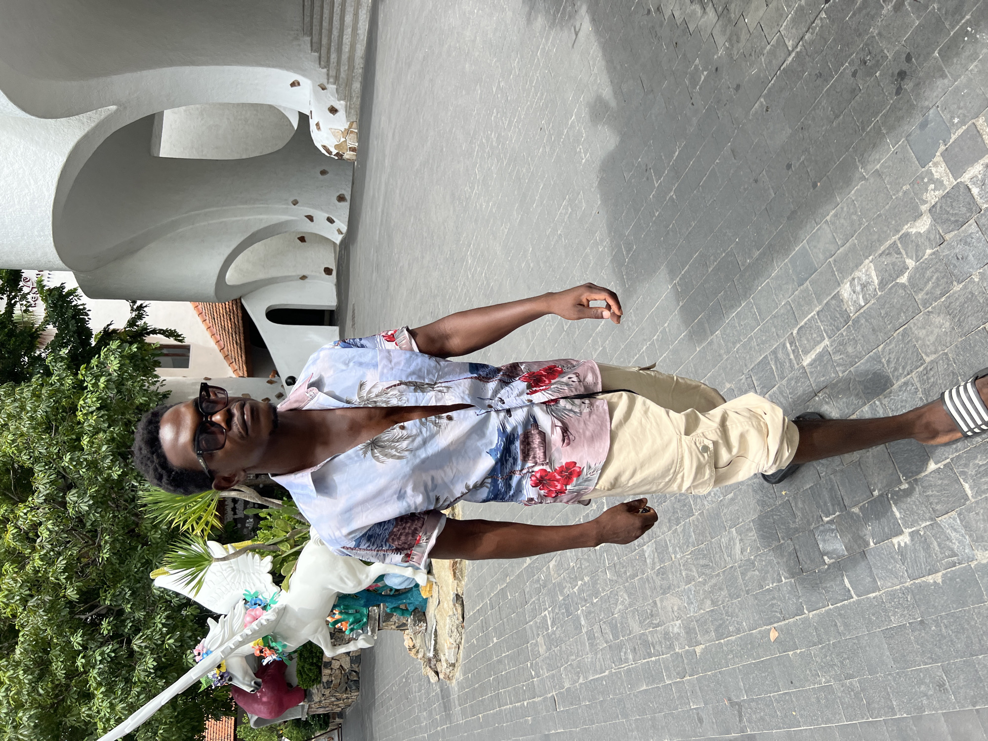 A dark-skinned man in sunglasses, a light-colored short-sleeved shirt with a floral and beach-themed print, and beige cargo shorts walks forward on a grey cobblestone path. He is on the right side of the frame, looking to his right. Behind him on the left is a white unicorn statue adorned with flowers, and behind the man on the right is a white, modern building with curved architectural elements. The ground is paved with grey cobblestones. The background shows lush green trees and a partially visible white building with an orange-tiled roof. The scene is lit by daylight, casting shadows on the ground. This is the entrance to the Khu Du Lich Truong Son.