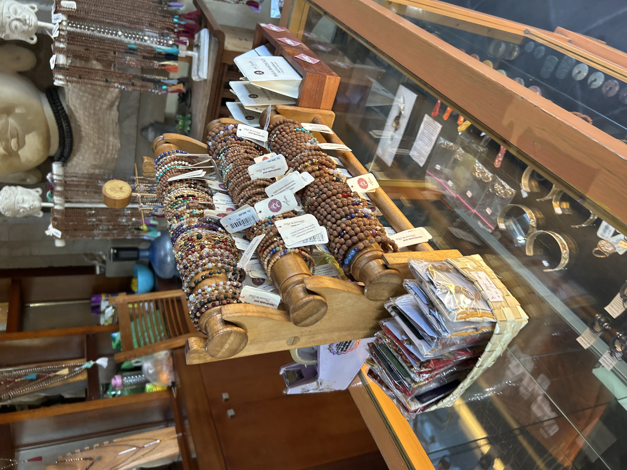 The image depicts a retail display of jewelry, likely in a shop located in Ubud District, Indonesia. The foreground is dominated by wooden stands holding numerous beaded bracelets, many with colorful beads and tassels. Several of these bracelets have white tags attached. To the left and slightly below the bracelet display, a woven basket holds several small, individually packaged items, possibly more jewelry or accessories. The background features a large, light-colored bust of a serene figure, possibly a Buddha statue, with smaller white figures on either side. Behind this, various strings of beads and decorative items are visible, along with wooden furniture and shelves. The overall impression is of a craft or spiritual goods store.