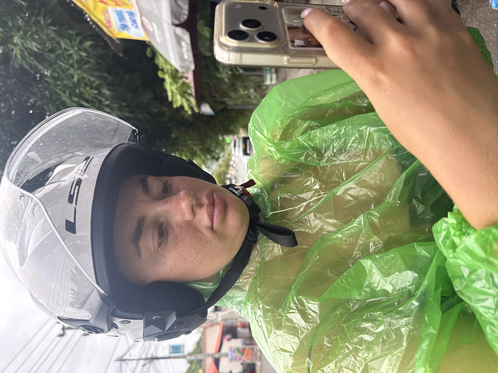An outdoor, medium shot, waist-up portrait shows a young Caucasian female wearing a white, full-face helmet with the visor up, and a bright green, translucent, plastic raincoat. She has fair skin with freckles, light brown hair partially visible under the helmet, and is looking down at a gold-colored smartphone in her right hand. The phone is held vertically and features a multi-lens camera system. Her left hand is positioned near the phone. The background suggests an urban street scene in Nha Trang, Vietnam, with blurred foliage, power lines, and a yellow sign with Vietnamese text. The overcast sky and the presence of water droplets on the helmet indicate it is raining.