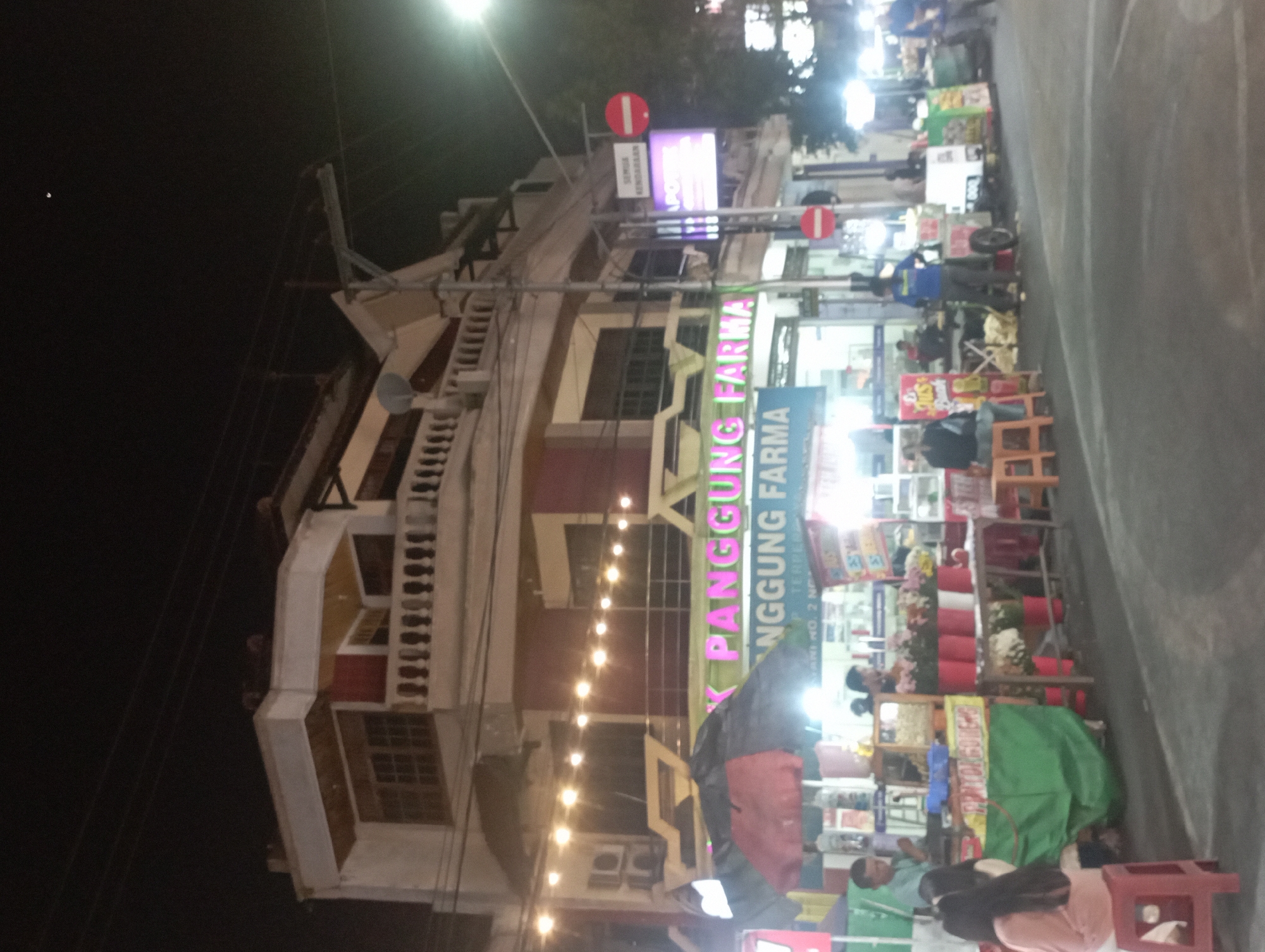 This is a nighttime street scene in Nganjuk, Indonesia, featuring a two-story building with a balcony and several illuminated signs. The building has a light-colored facade and a dark-colored roof. In front of the building are several market stalls or food vendors. One stall has a green canopy and displays various items, including flowers in red and white buckets on a table. Another stall is partially visible with a pink and black umbrella.
Several people are present. On the left, a person in a light blue shirt stands behind a stall. To their right, a woman in a pink top is seated on a red stool. In the center, a person wearing a dark shirt is seated on an orange stool. To the right of the center, a person in a blue shirt stands near a cart. Further to the right, another person in a blue shirt is visible. In the background, a few more individuals are scattered among the stalls.
Illuminated signs are prominent, including one in pink neon that reads "PANGGUNG FARMA". Another sign above it is a purple illuminated rectangular sign with text. Two red circular signs with white horizontal bars are also visible on poles, indicating a prohibition. String lights are strung across the scene, adding to the nighttime ambiance. The street is paved, and the overall impression is of a lively nighttime marketplace.