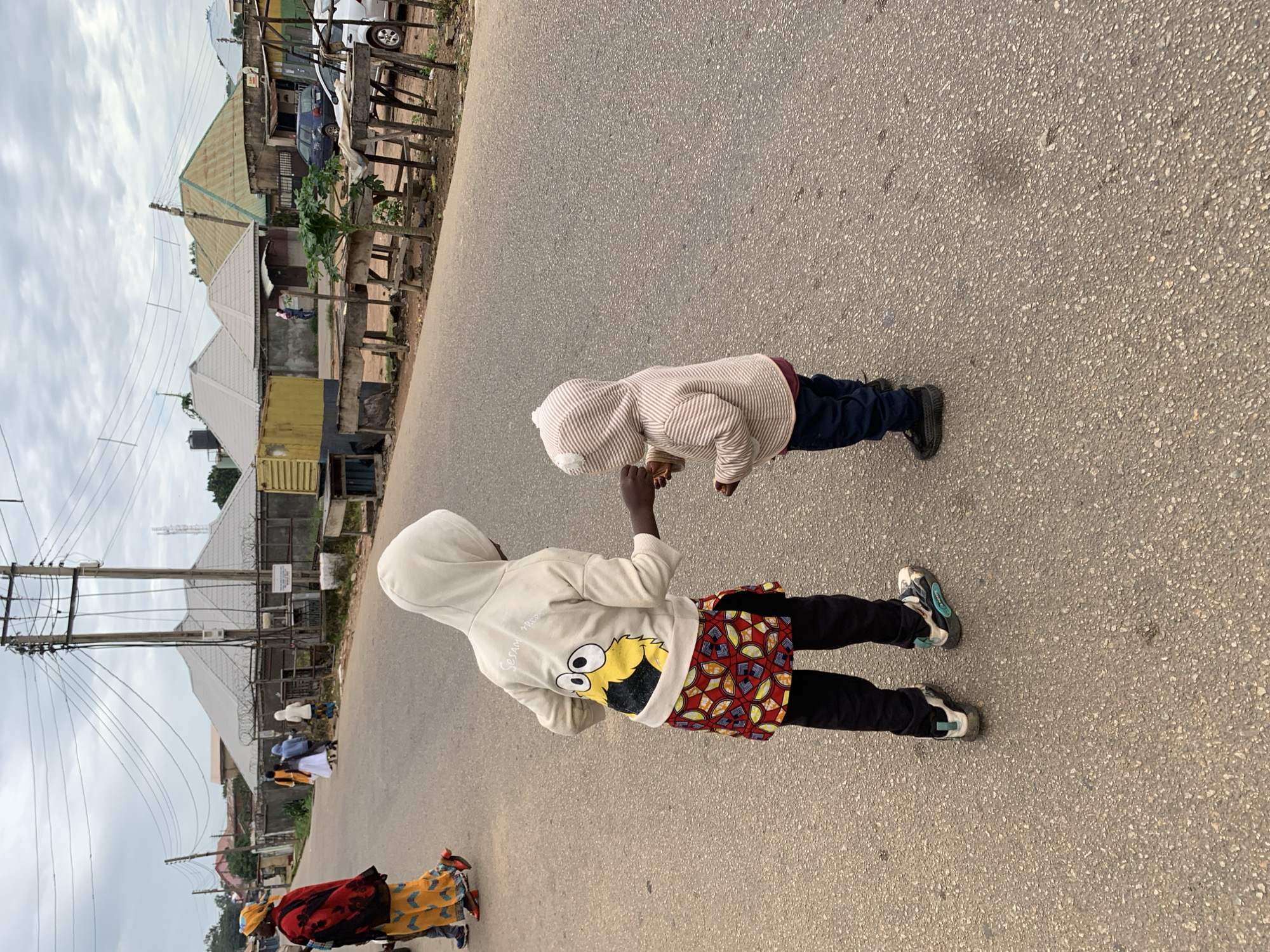 Two children, one older and one younger, stand on a paved road in Jos, Nigeria. The older child, facing away from the camera, wears a light-colored hooded sweatshirt with a cartoon character on the back and a colorful patterned skirt over black pants. They are handing something to the younger child, who is also facing away and wearing a striped hooded sweatshirt and dark pants. In the background, other people walk along the road, and various buildings and market stalls are visible. Utility poles with numerous wires are prominent in the sky. The overall environment appears to be an urban street scene under an overcast sky.