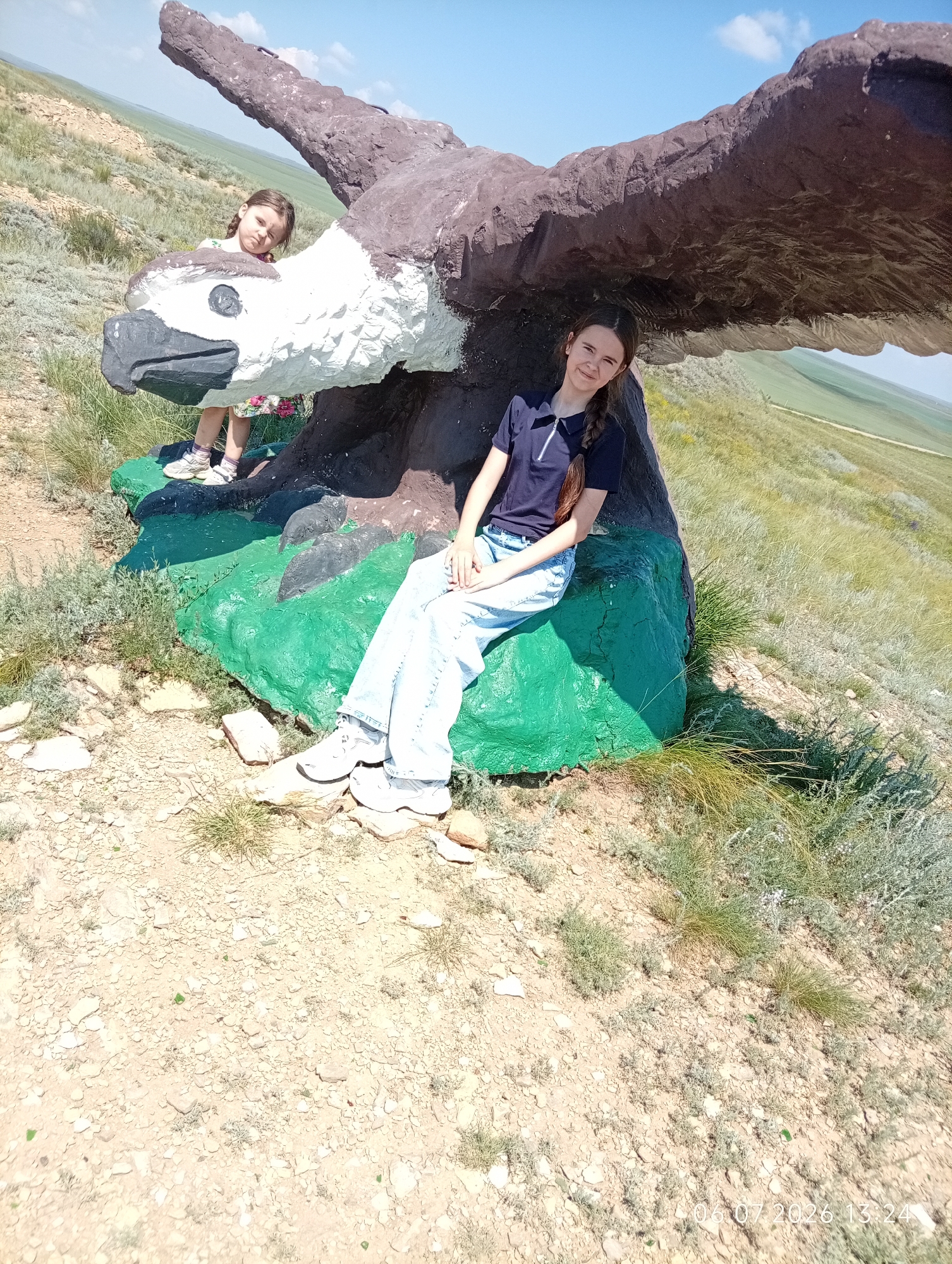 In a landscape with rolling hills and dry vegetation under a clear blue sky, two young females are photographed next to a large, concrete sculpture of an eagle. The sculpture is predominantly brown and white, with green painted rocks at its base. The elder female, with long brown hair braided, is seated on the green rocks beneath the eagle's outstretched wing. She is wearing a dark blue polo shirt and light-wash denim jeans. Her posture is relaxed, with her arms resting on her lap. Behind her and to the left, a younger female, with lighter brown hair, is standing, partially obscured by the eagle's head and body. She is wearing a patterned dress and white sneakers. The ground is covered with dry earth, small stones, and sparse grasses. The lower right corner of the image displays a timestamp: "06.07.2026 13:24". The scene appears to be outdoors in a rural or semi-natural environment, possibly in the vicinity of Aleksandrovka, Kazakhstan, based on the geological features and vegetation.