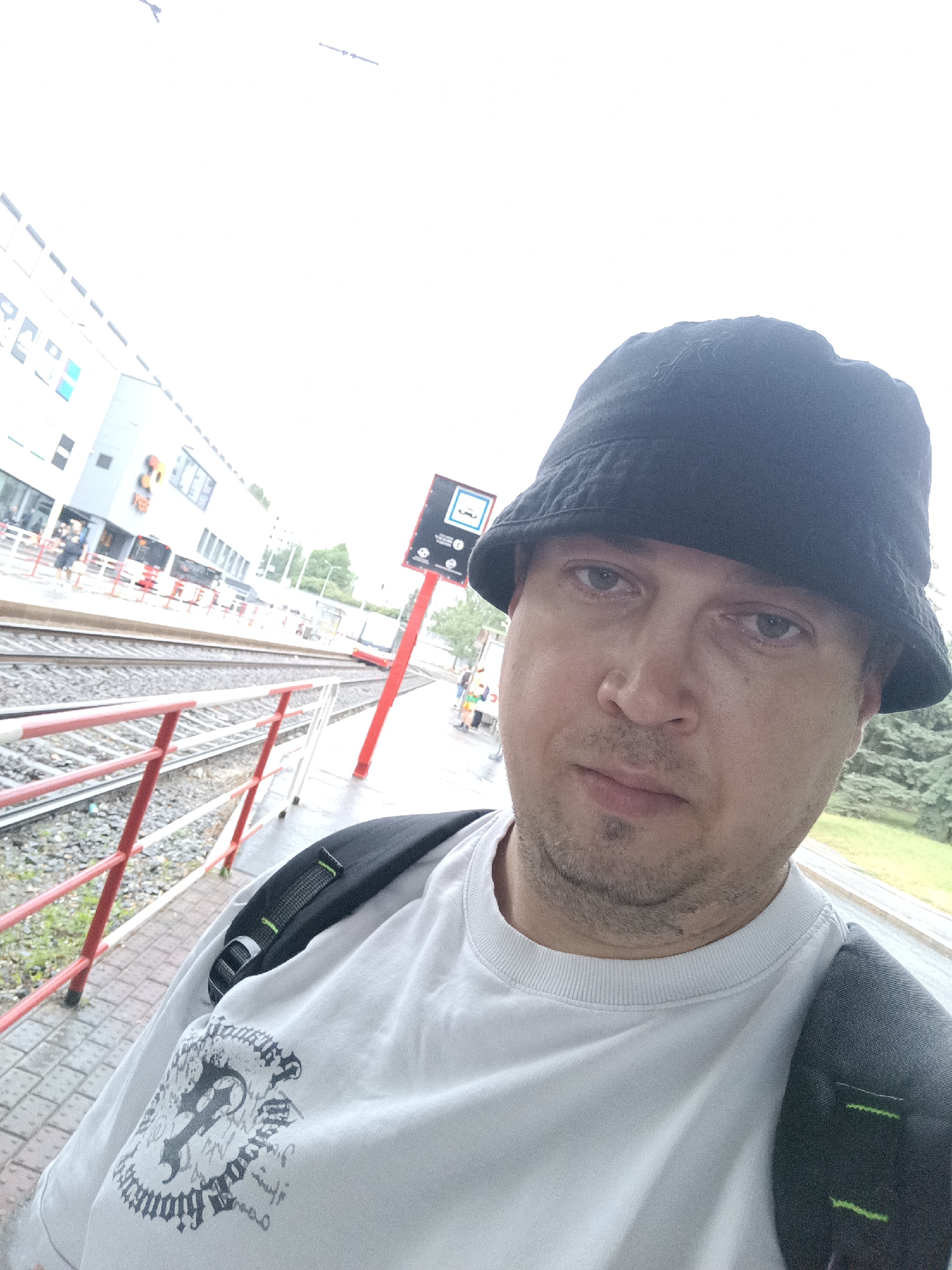A man wearing a black bucket hat and a white sweatshirt takes a selfie at a tram station in Prague, Czechia. He has a backpack on and is standing on a wet platform. In the background, there is a tram, buildings, trees, and railway tracks. The weather appears to be overcast.