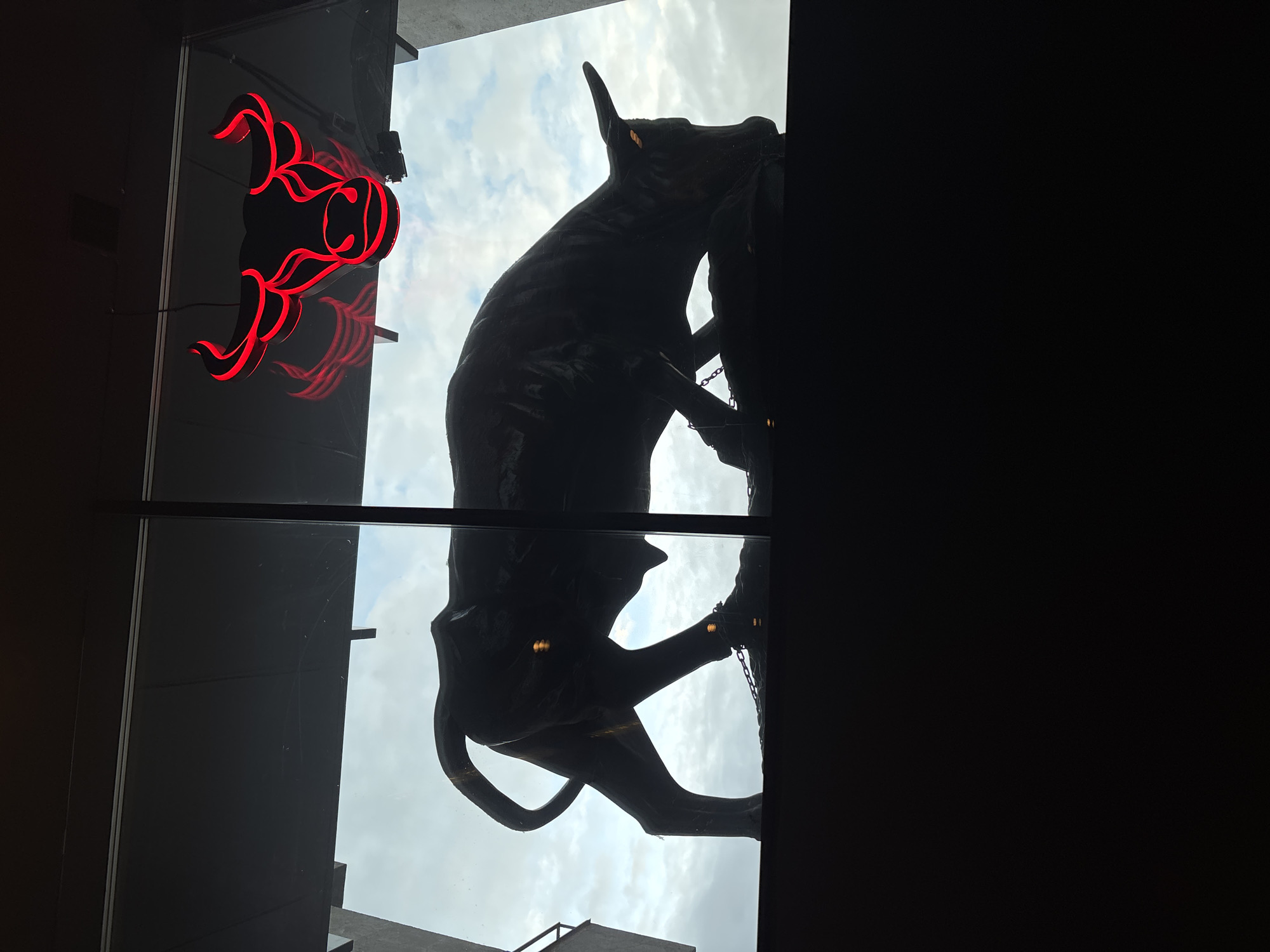 The image shows the exterior of a building, likely a restaurant or establishment, in Moscow, Russia. A large silhouette of a bull statue, seemingly made of metal, is seen, as well as a decorative red neon sign of what seems to be a bull's head above. The view is through a window looking upwards, with the sky and some cloud cover visible in the background. The scene appears to be from an indoor perspective, looking out at the exterior elements.