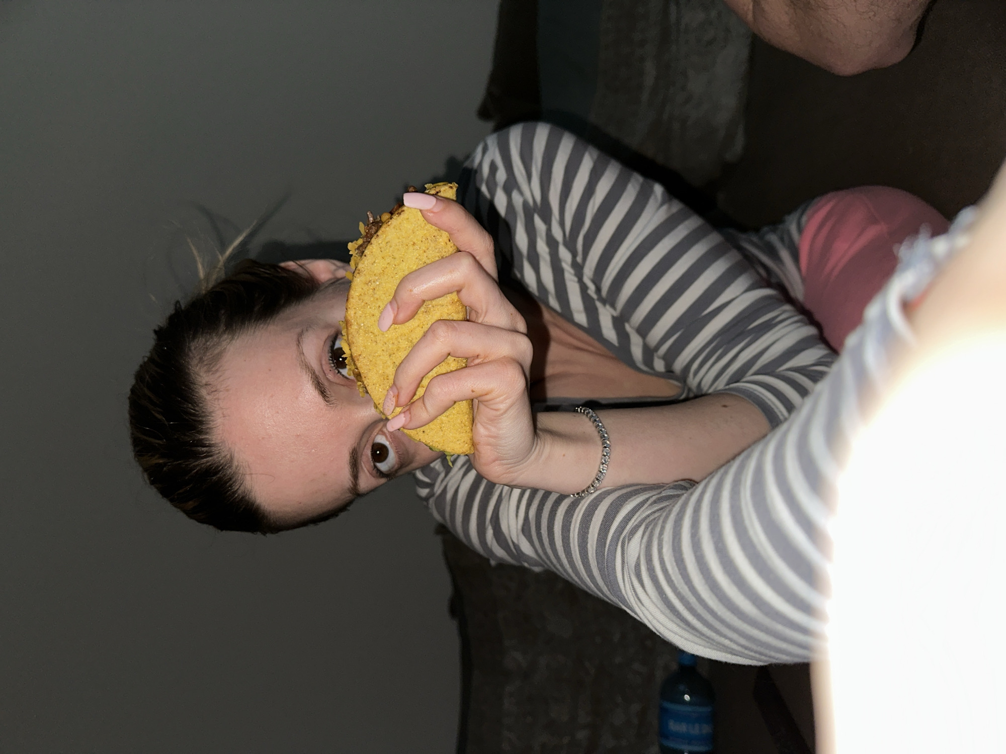 A candid, rotated portrait shows a person holding a hard-shell taco close to their face, looking directly at the viewer with wide, dark eyes. The person, with dark hair pulled back, occupies the left and central parts of the frame, tilted on their side. They are wearing a grey and white striped long-sleeved top, with a hint of a pink garment underneath. A silver-toned bracelet is visible on their right wrist, and their fingernails are painted a light pink. The taco appears to be filled with ground meat and cheese, some of which is visible at the top. The background is dark and indistinct, suggesting an indoor, dimly lit setting. A strong, bright light source emanating from the right side of the image causes a significant lens flare, partially obscuring the right edge. In the lower right corner, a blue bottle, possibly containing water or soda, is partially visible, with blurred text "SAN LO" discernible on its label. The scene takes place in Amsterdam, Netherlands.