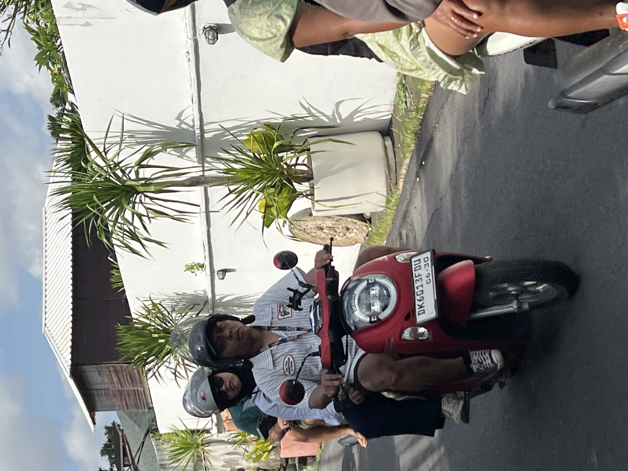 A man and two passengers are riding on a red scooter on a paved street in North Kuta, Indonesia. The man in the front is wearing a striped shirt and a black helmet. The passenger behind him is wearing a silver helmet and sunglasses. The third passenger, on the far back, is partially visible and is wearing a green shirt. The scooter has a license plate that reads "DK6G13FDU 06-30". Behind them is a white wall with plants in pots and a building with a corrugated metal roof. The sky is blue with some clouds.