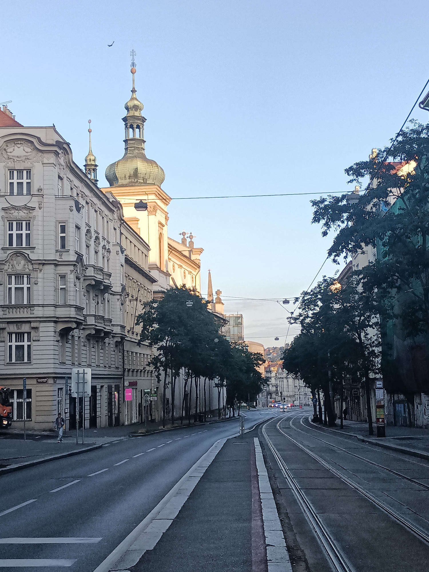 The image depicts a street scene in Prague, Czechia, likely during the early morning or late evening due to the soft lighting and clear sky. The perspective is from a street level, looking down a road with tram tracks running along the right side of the frame.
On the left side of the street, there is an ornate, multi-story building with classical architectural features, including detailed facades, balconies, and numerous windows. The building appears to be a mix of residential and commercial spaces, with signage visible at street level, indicating shops or businesses. A pedestrian can be seen walking on the sidewalk near a crosswalk.
To the right of the classical building, a prominent church or cathedral structure rises, characterized by its distinctive green-domed tower topped with a cross, and a smaller, similar tower.
The right side of the street is lined with trees, casting shadows on the road and sidewalk. Behind the trees, more buildings are visible, some appearing to be older structures with historical architecture, while others seem more modern.
The road itself is paved and shows clear lane markings and tram tracks. In the distance, more of the city unfolds, with buildings and a hint of hilly terrain. The sky is a pale blue, with no clouds visible, and a single bird is captured in flight high above the buildings. The overall atmosphere is calm and quiet, suggesting a less busy time of day in the city.