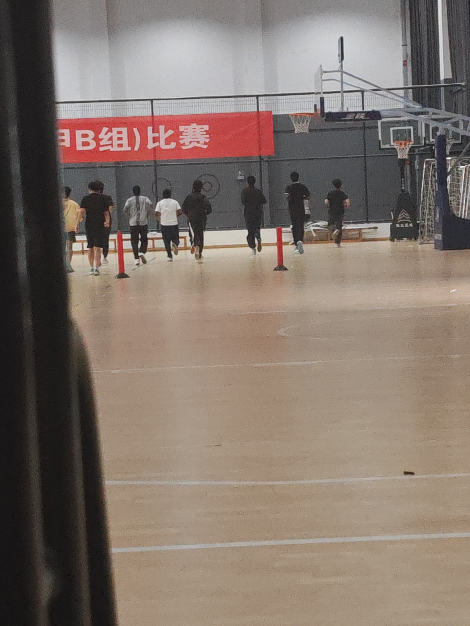 A low-resolution image shows several people running on a basketball court in what appears to be a gymnasium in Jiangning District, China. The people are all facing away from the camera, and they are running toward the back of the gym. There is a red banner hanging on the wall behind them with Chinese characters. There are basketball hoops visible, and the floor is marked with white lines. A dark, blurred object is in the foreground on the left side of the image. The overall lighting is bright, and the scene seems to be a training or practice session.