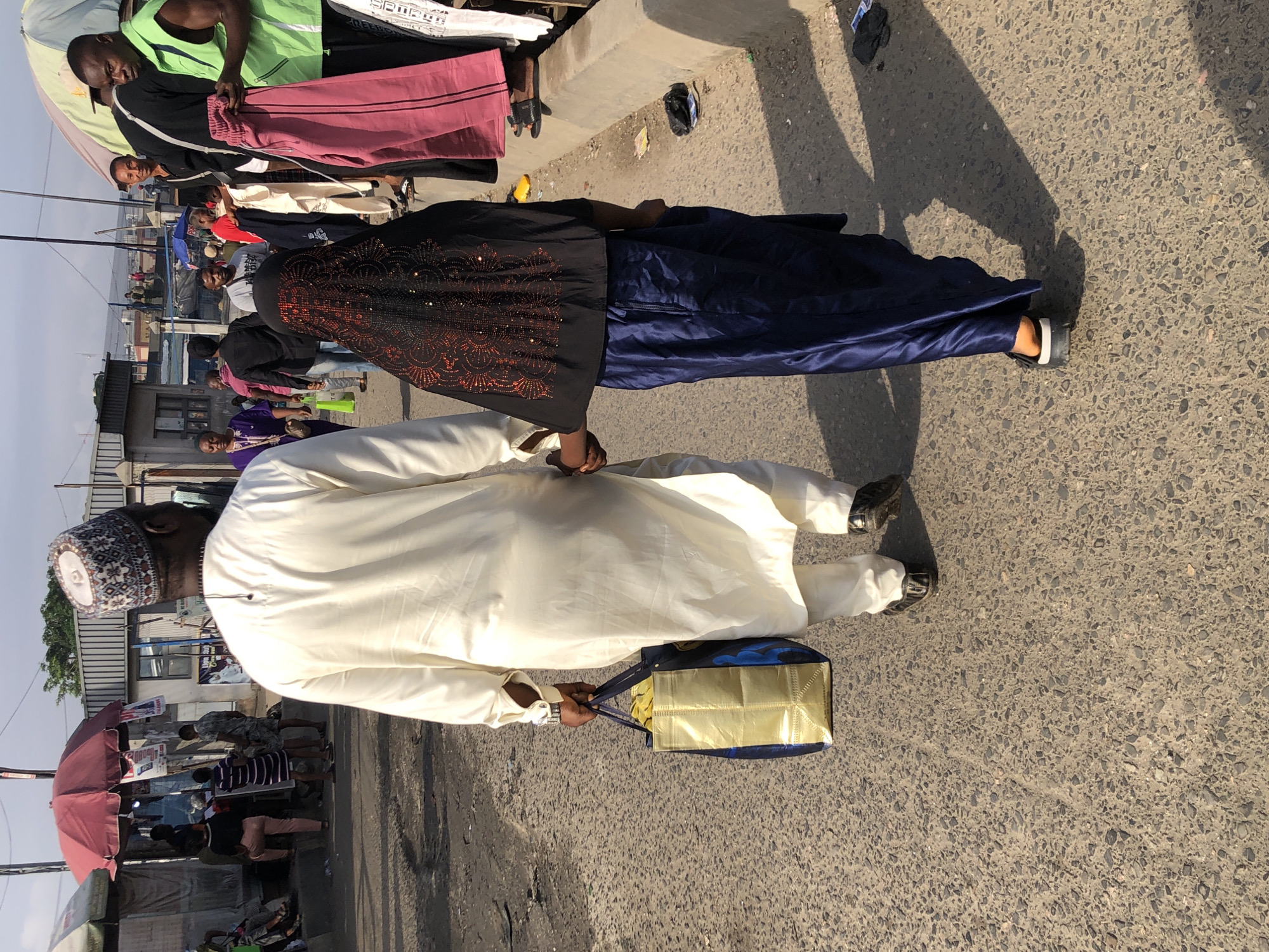 A man and a young girl walk hand-in-hand on a paved street in Lagos, Nigeria. The man, from behind, wears a white traditional robe and a patterned cap, holding a gold and blue patterned shopping bag in his left hand. The girl, also viewed from behind, wears a black hijab with red sequin embellishments and a flowing navy blue skirt. They are walking away from the camera in a bustling outdoor market environment. Stalls and other people are visible in the background, suggesting a busy commercial area. The sun casts long shadows on the ground, indicating it is likely late afternoon.