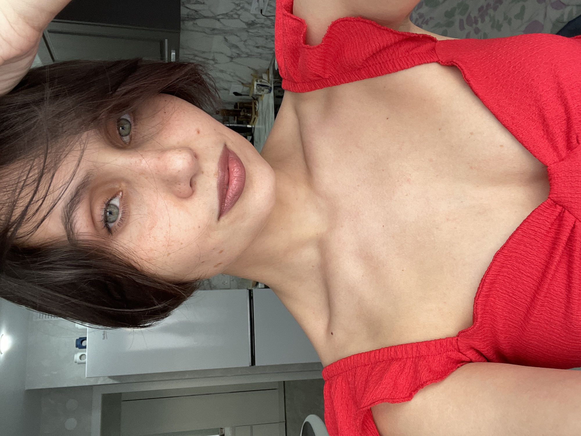 A woman with short brown hair and green eyes is facing the camera, wearing a red top. She appears to be indoors, with a kitchen in the background featuring white cabinets and a marble countertop. The location in Izhevsk, Russia, cannot be determined from the visual information provided.
