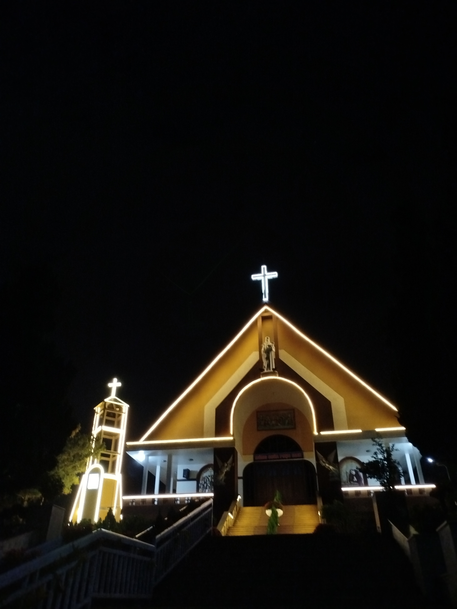 The image shows a brightly illuminated church at night. The church features a central structure with a triangular roof, a prominent cross atop it, and a statue below. The building is trimmed with yellow lights outlining its shape. To the left is a bell tower, also illuminated. A set of stairs leads up to the entrance. The location is in Lạc Dương, Vietnam.