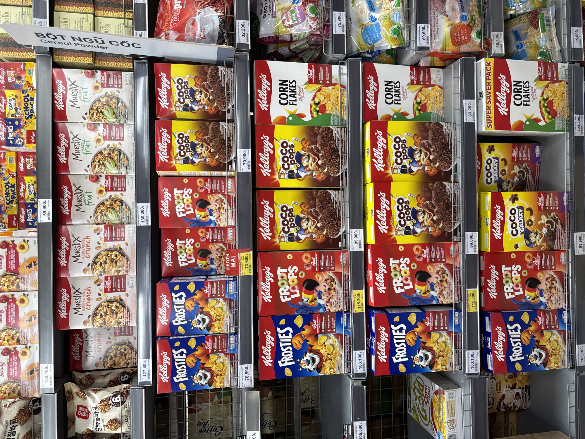 The image displays a retail store shelf in Nha Trang, Vietnam, filled with various Kellogg's cereal boxes. The products are neatly arranged on multiple tiers of metal shelving. Prominently featured are Kellogg's Frosties, Froot Loops, Coco Pops, Mueslix, and Corn Flakes, some of which have Vietnamese labeling. Prices are visible on some of the shelves, indicated by small labels with currency. In the background, other grocery items are visible, suggesting a supermarket or convenience store setting. The lighting appears to be overhead store lighting, illuminating the products clearly. There are no people visible in the image.