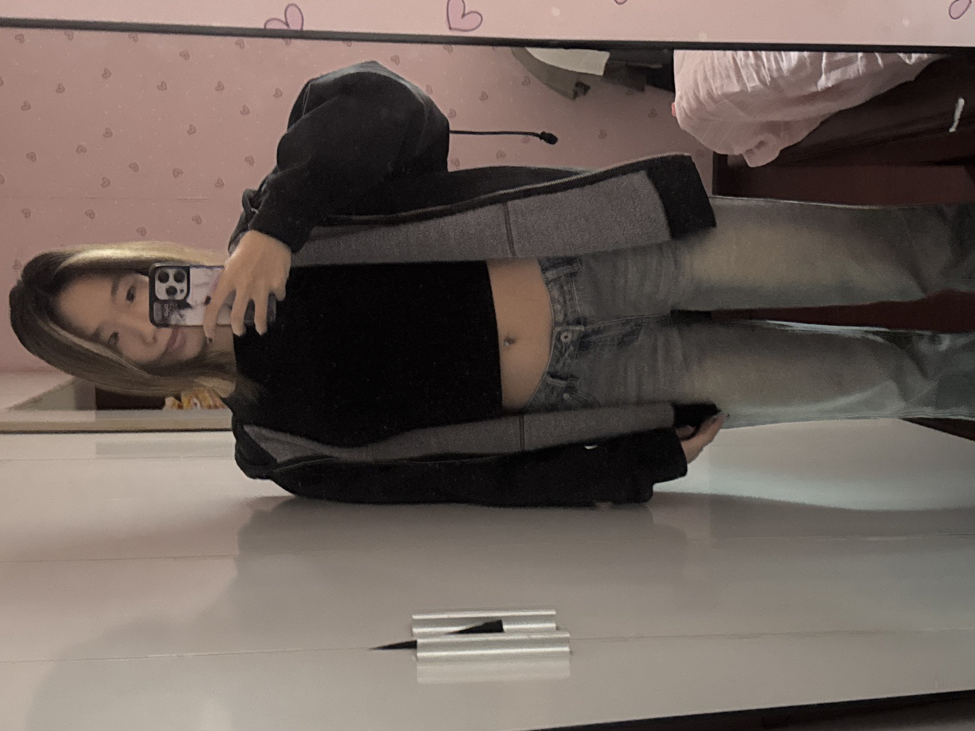 A person is visible in a mirror reflection. The person is holding a cell phone in their hand and appearing to take a picture of themselves. They are wearing a black top and jeans. The background features a wall with heart pattern. The reflection suggests the setting might be in Shapingba District, China, but this is based on limited visual information.